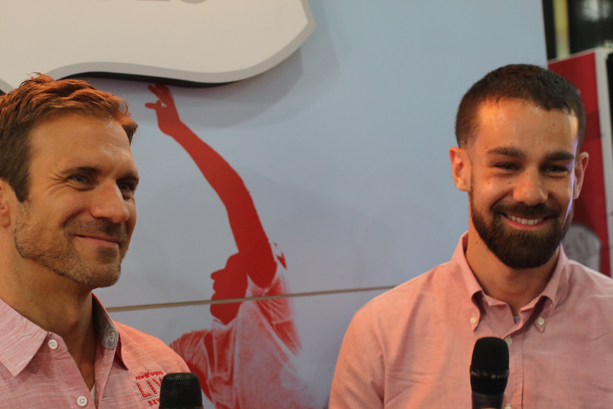 Two smiling men in pink shirts with microphones at the bottom of the frame, in front of a pale blue backdrop with a red graphic.