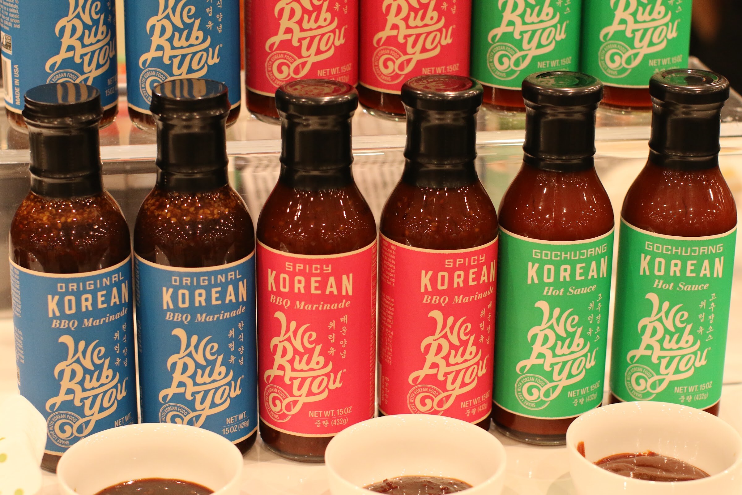 Six bottles of We Rub You sauces with blue, red and green labels above three bowls of sauce.