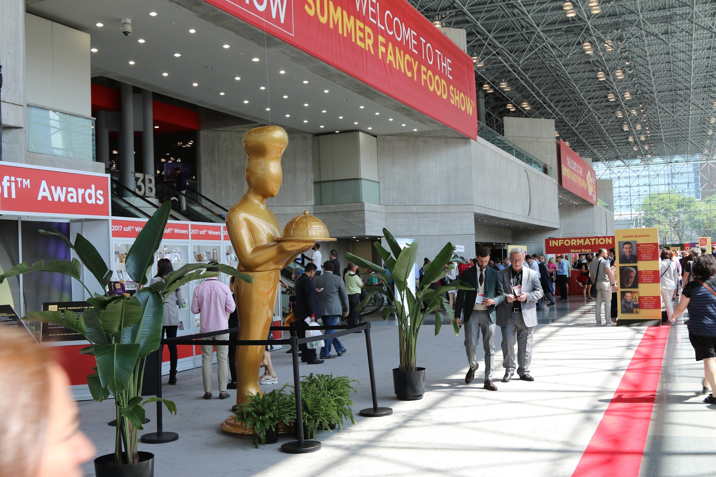 A red Summer Fancy Food Show banner over a Javits Center entrance, with a gold chef statue holding a cloche beside a sofi Awards display and attendees walking past.