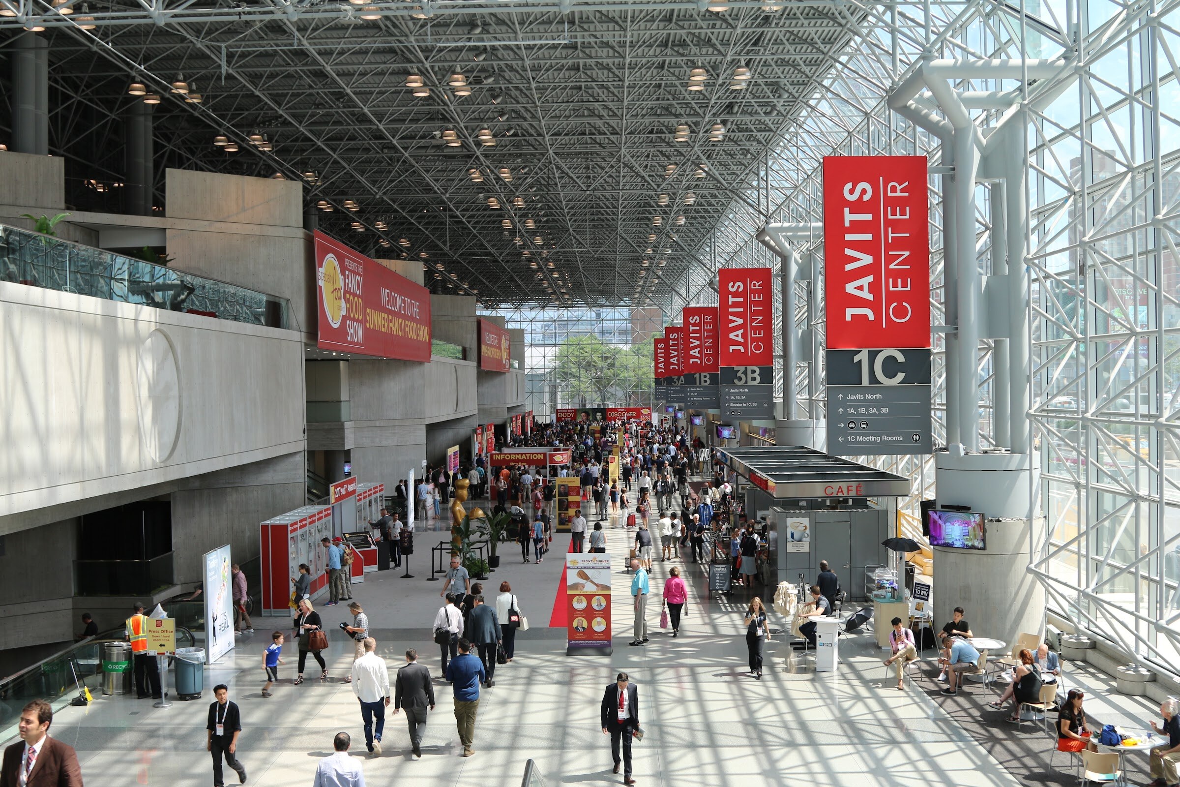 A long, sunlit convention concourse with Javits Center signs, Fancy Food Show banners and many attendees walking toward the exhibit halls.