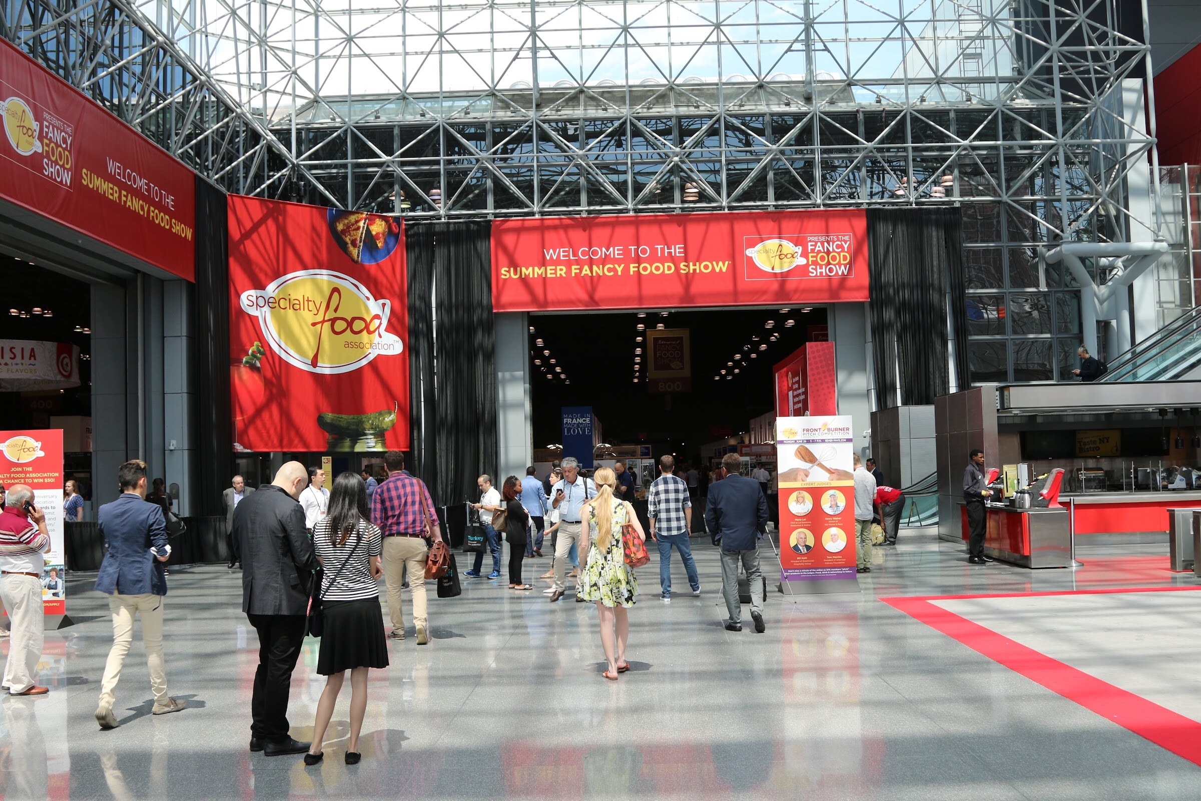 People walking toward a dark exhibit hall entrance beneath a Welcome to the Summer Fancy Food Show banner and a Specialty Food Association flag.