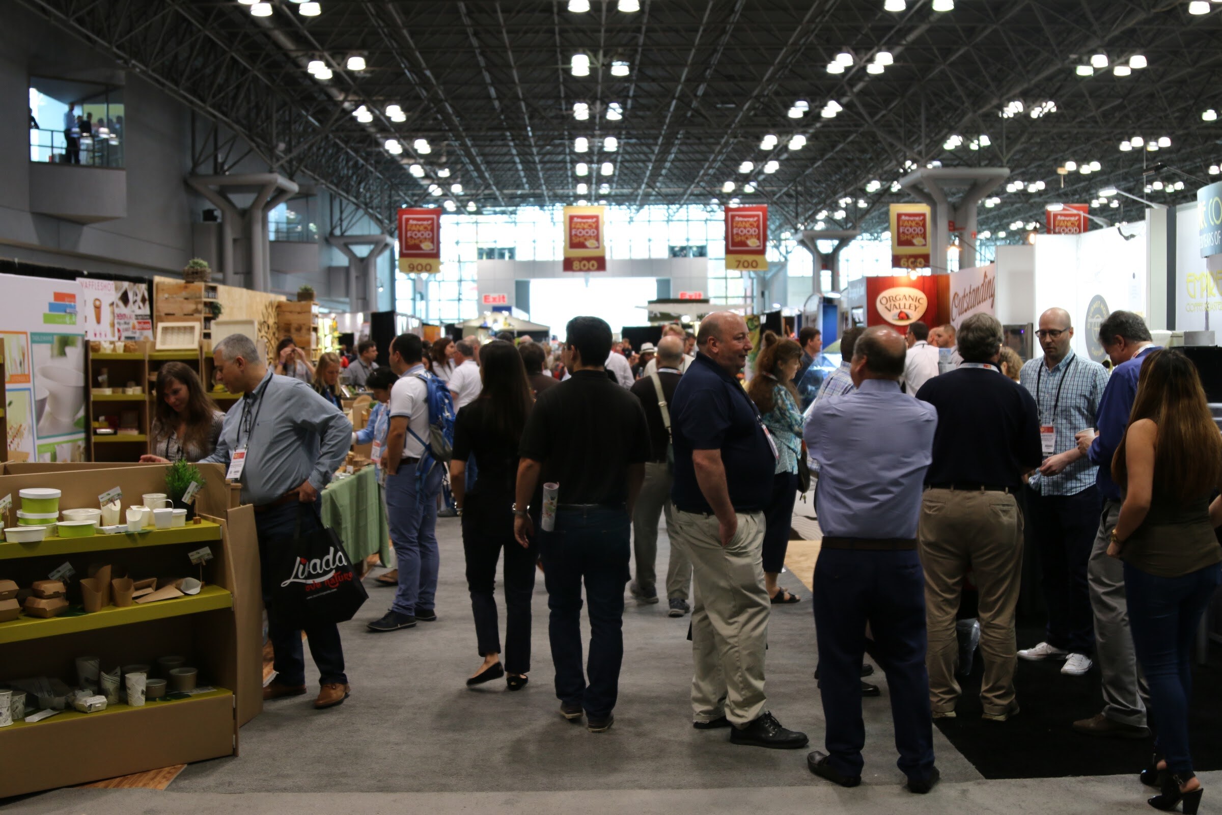 A crowd of badge-wearing attendees standing in a trade show aisle under hanging aisle number signs.