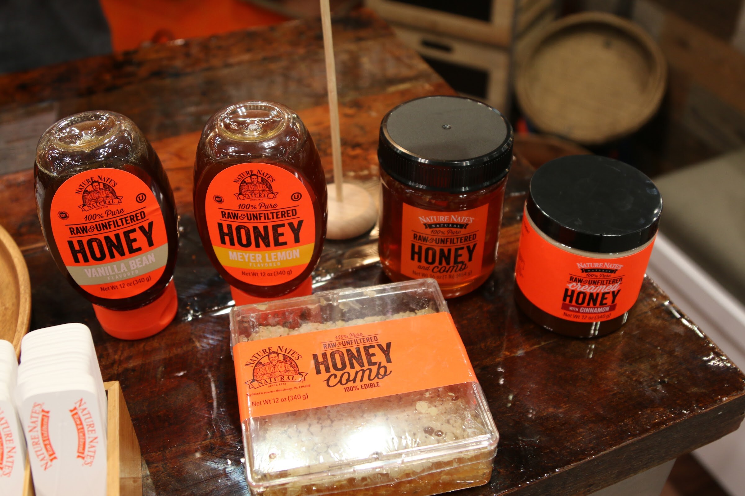 Orange-labeled Nature Nate's honey squeeze bottles, jars and a honeycomb box on a dark wooden table.