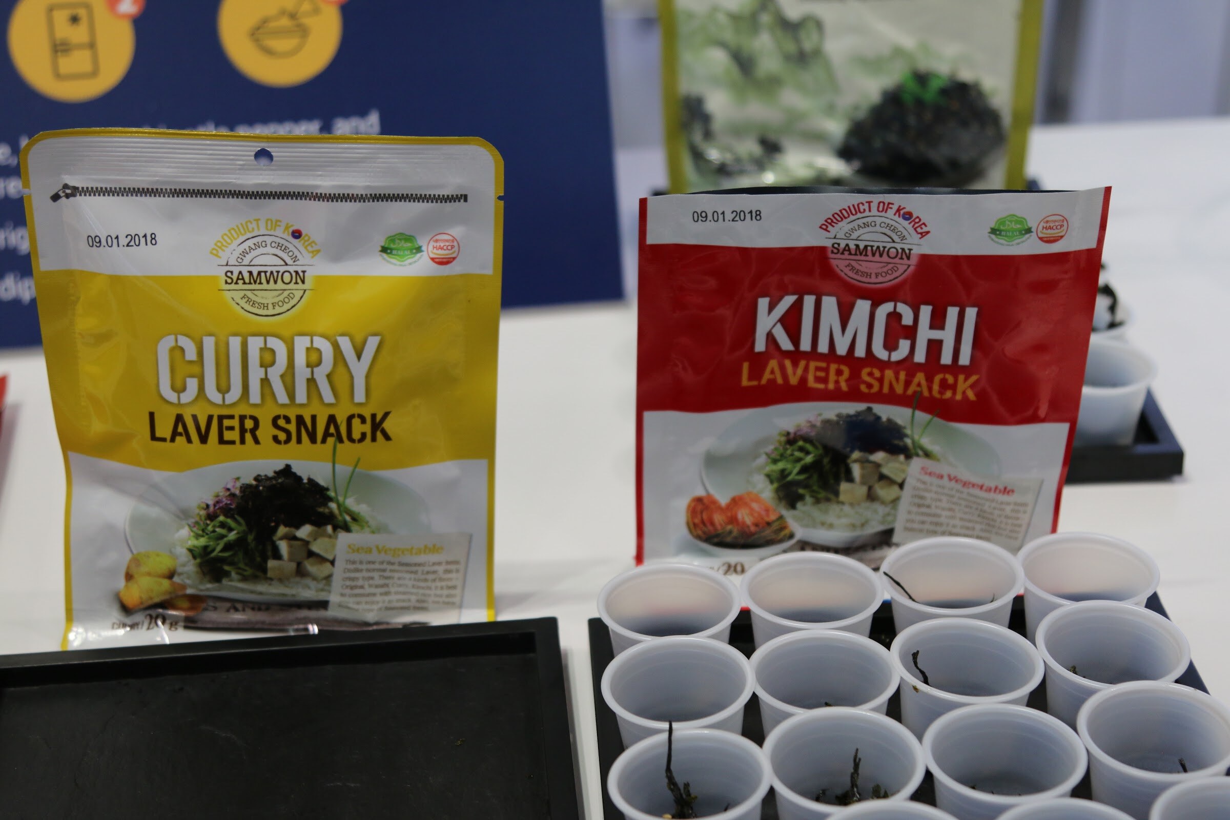 Two bags of laver snack, yellow curry and red kimchi, standing above a tray of empty plastic sample cups.