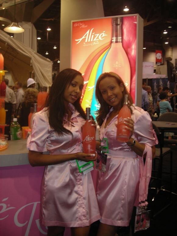 Two women in pink satin outfits hold bottles of Alizé in front of a lit Alizé sign at a trade show booth with a pink counter.