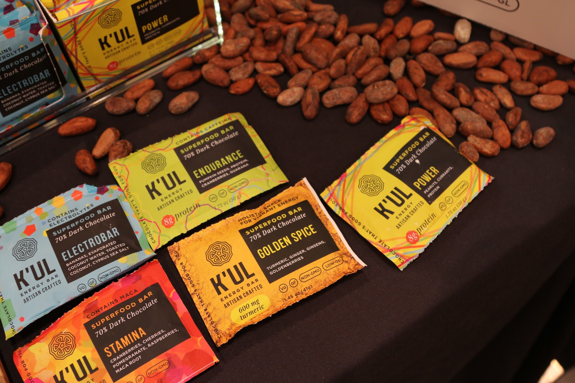 Colorful K'UL energy bar packages on a black table scattered with cacao beans.