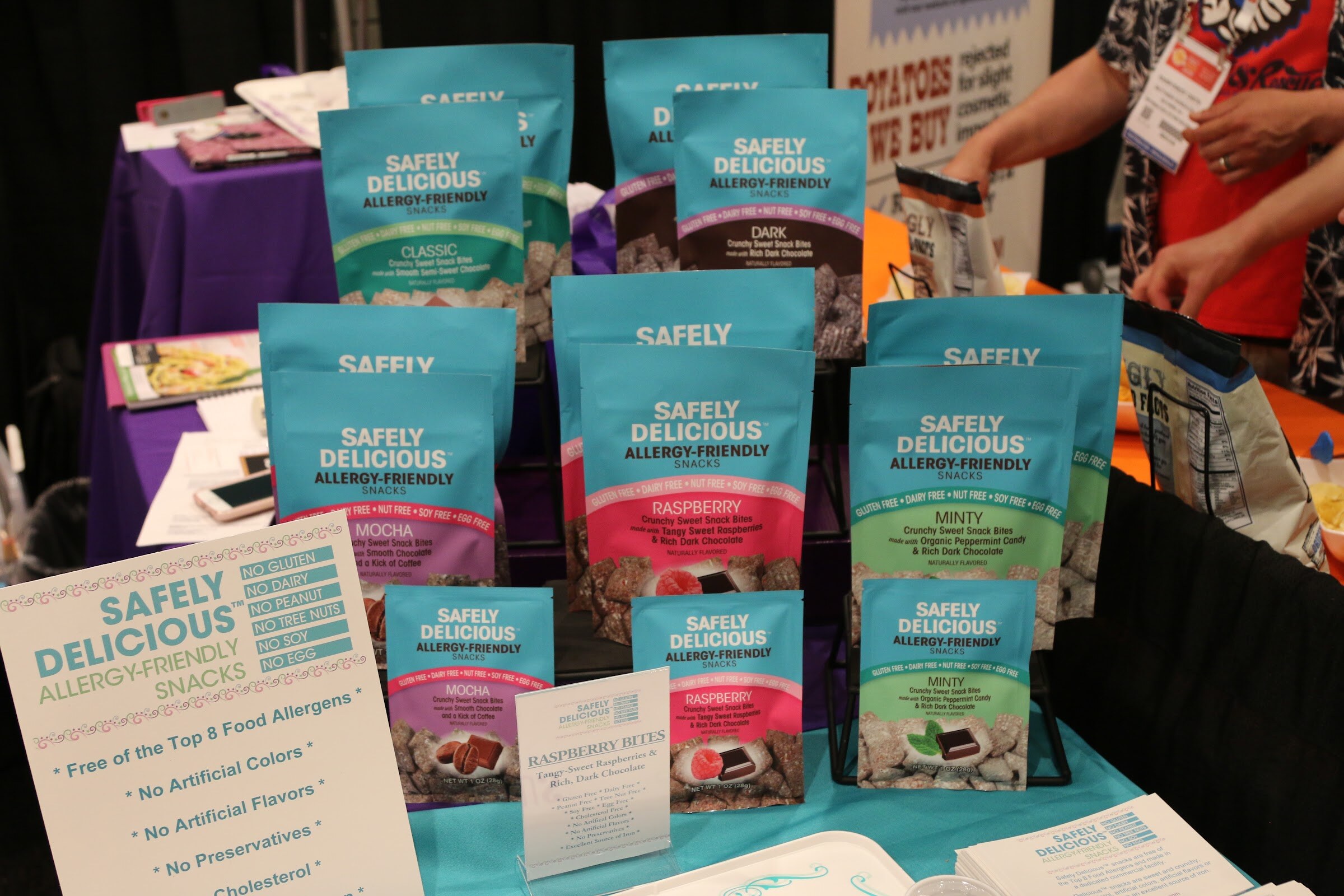 Teal Safely Delicious snack bags stacked on a rack with a sign about allergy-friendly snacks at the front.