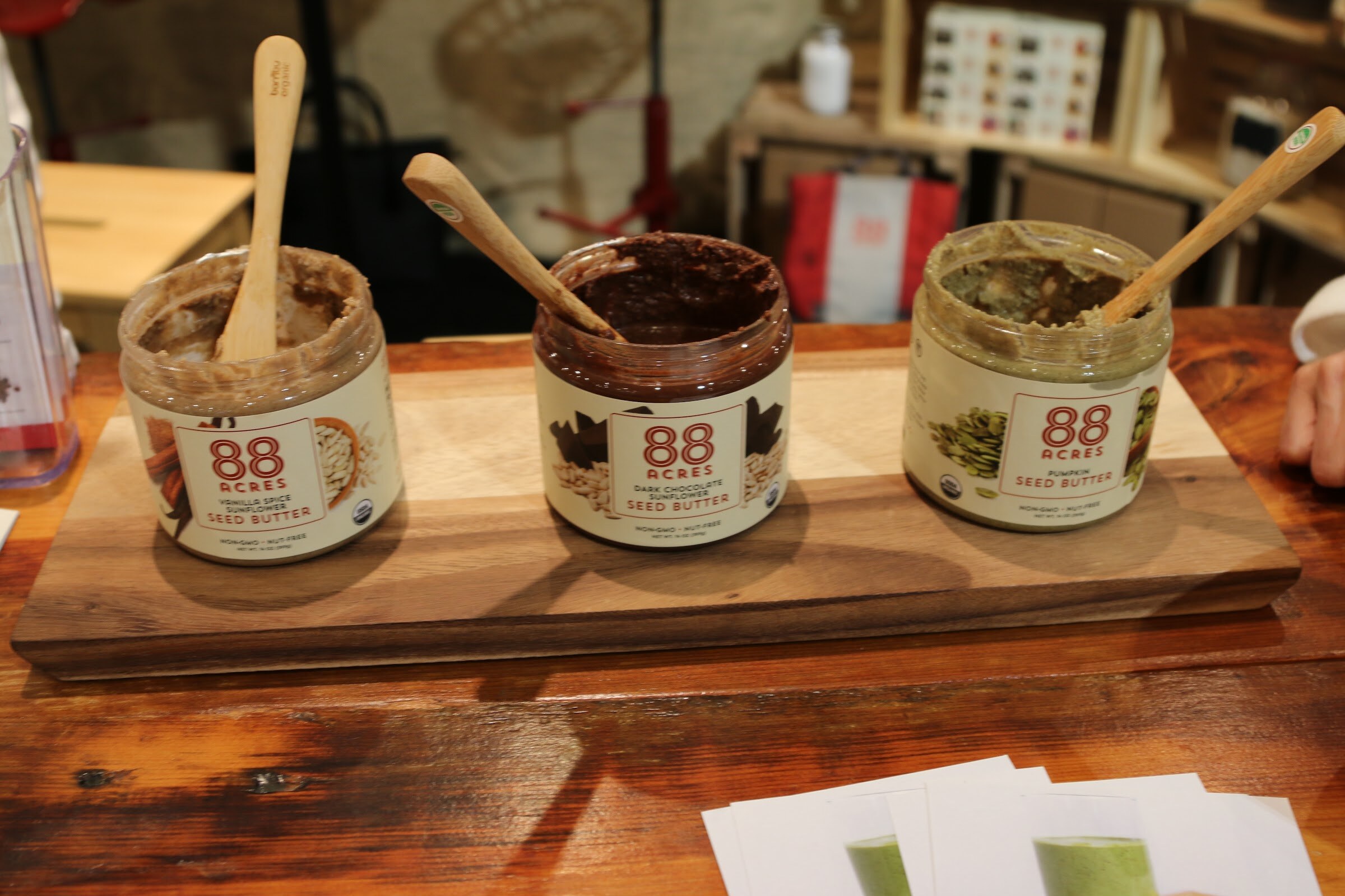Three open jars of 88 Acres seed butter with wooden spoons on a board.