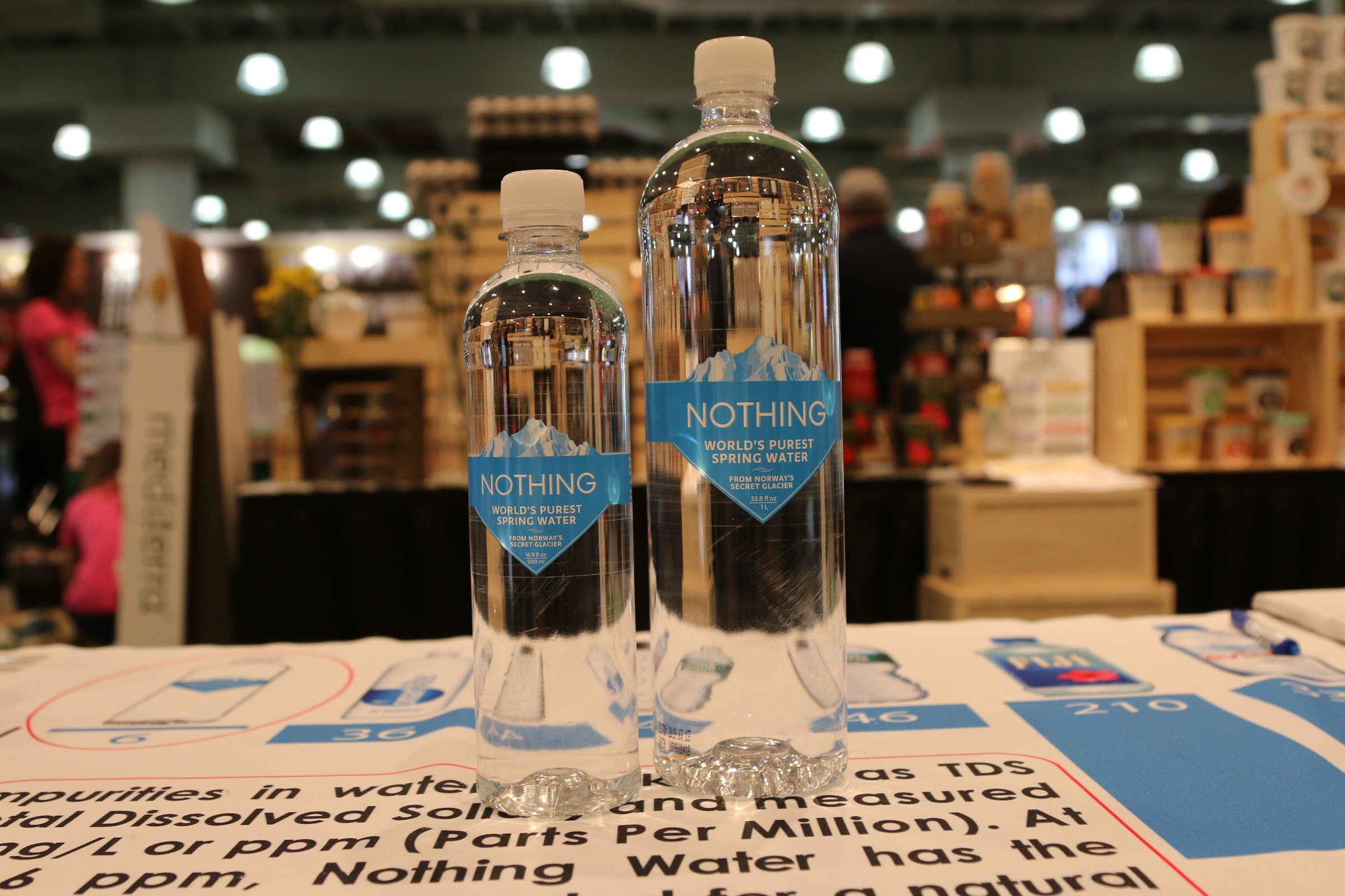 Two clear Nothing spring water bottles on a table with a printed chart, with the exhibit hall blurred behind.