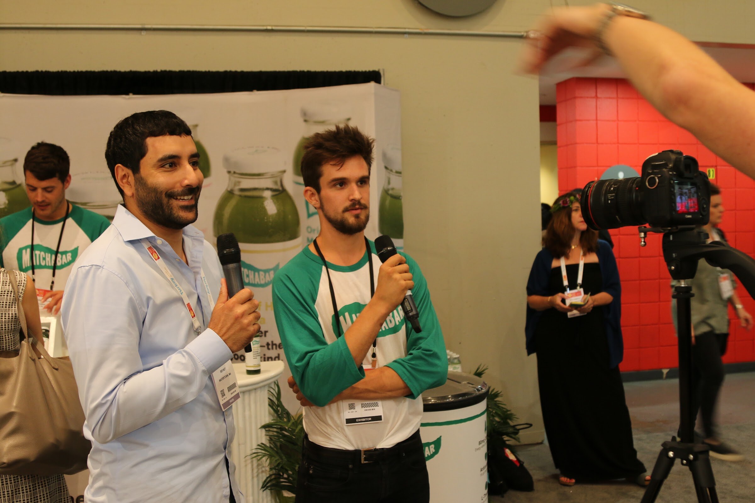 Two men in MatchaBar clothing holding microphones in front of a MatchaBar banner, with a video camera on a tripod at right.