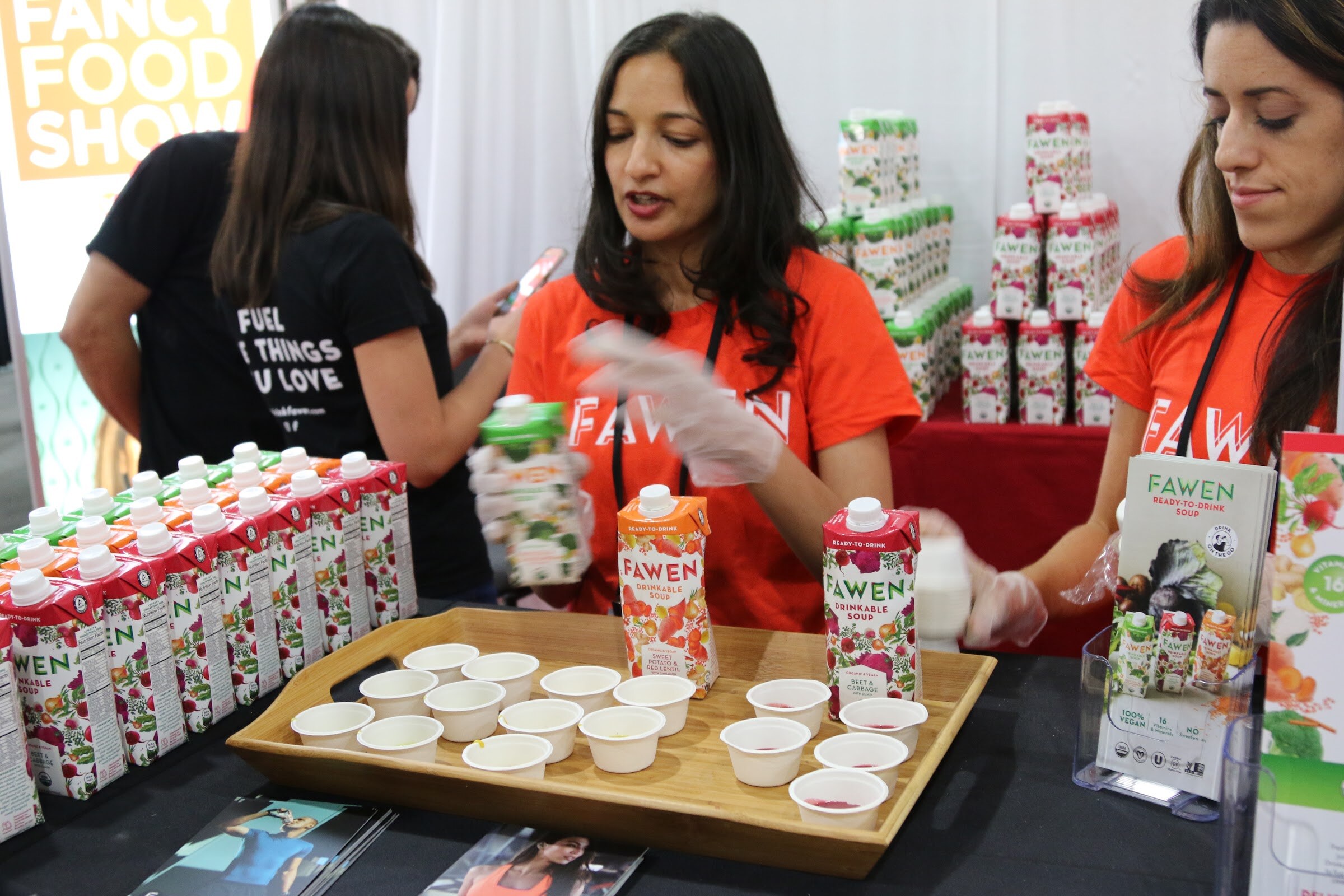 Two women in orange Fawen shirts preparing sample cups on a tray beside Fawen drinkable soup cartons.