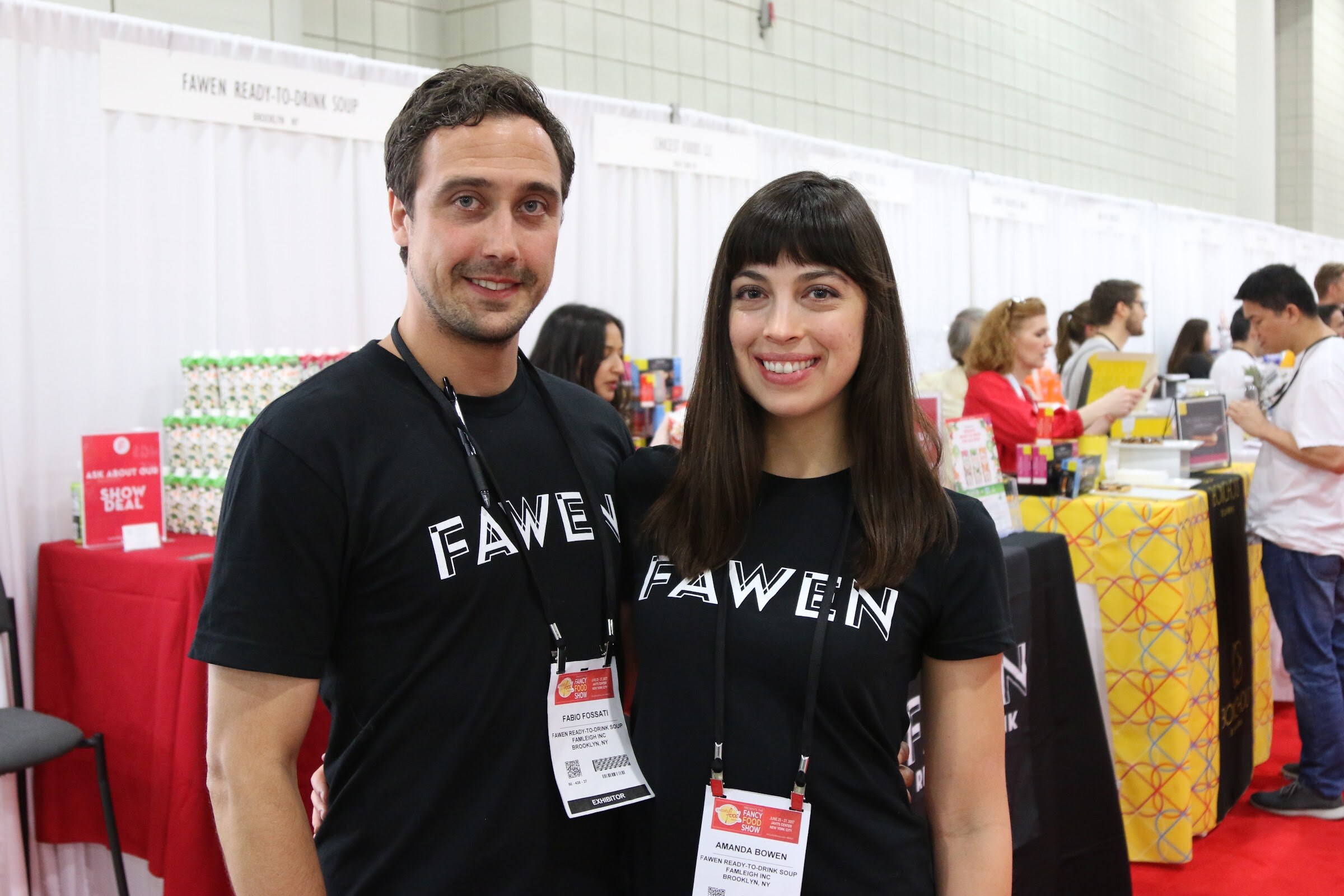 A smiling man and woman in black Fawen t-shirts with exhibitor badges standing in front of a booth with stacked cartons.