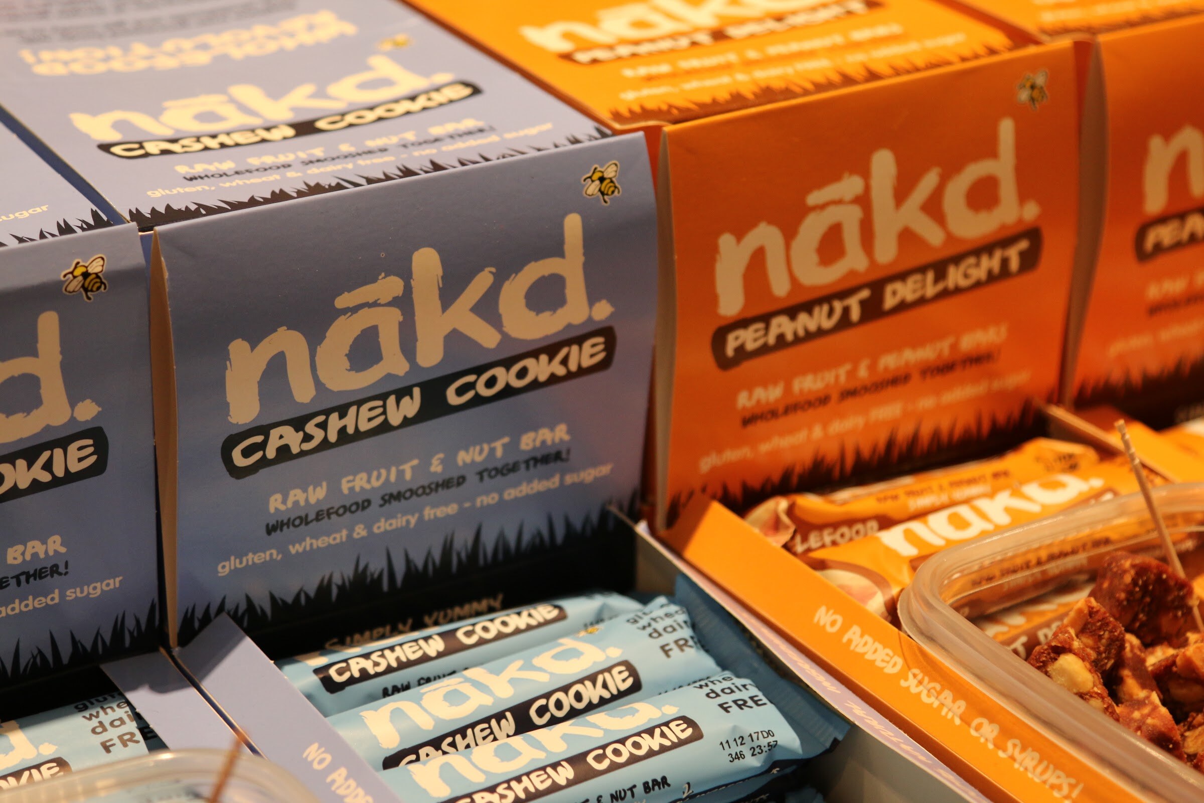 Blue nakd. Cashew Cookie and orange Peanut Delight display boxes with bars in front.