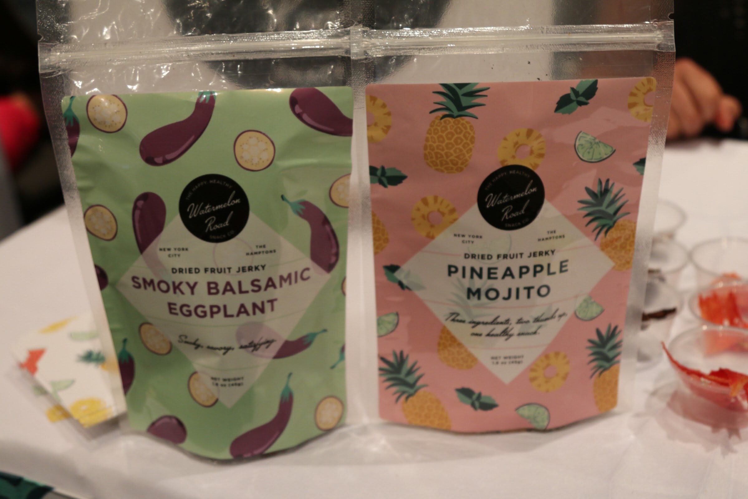 Two clear pouches of Watermelon Road dried fruit jerky, one green with eggplants and one pink with pineapples.