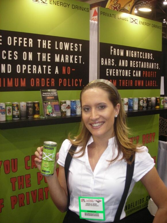 Angela Wise holds a can of X energy drink in front of Brand X Fluids private label energy drink signs and a shelf of assorted cans.