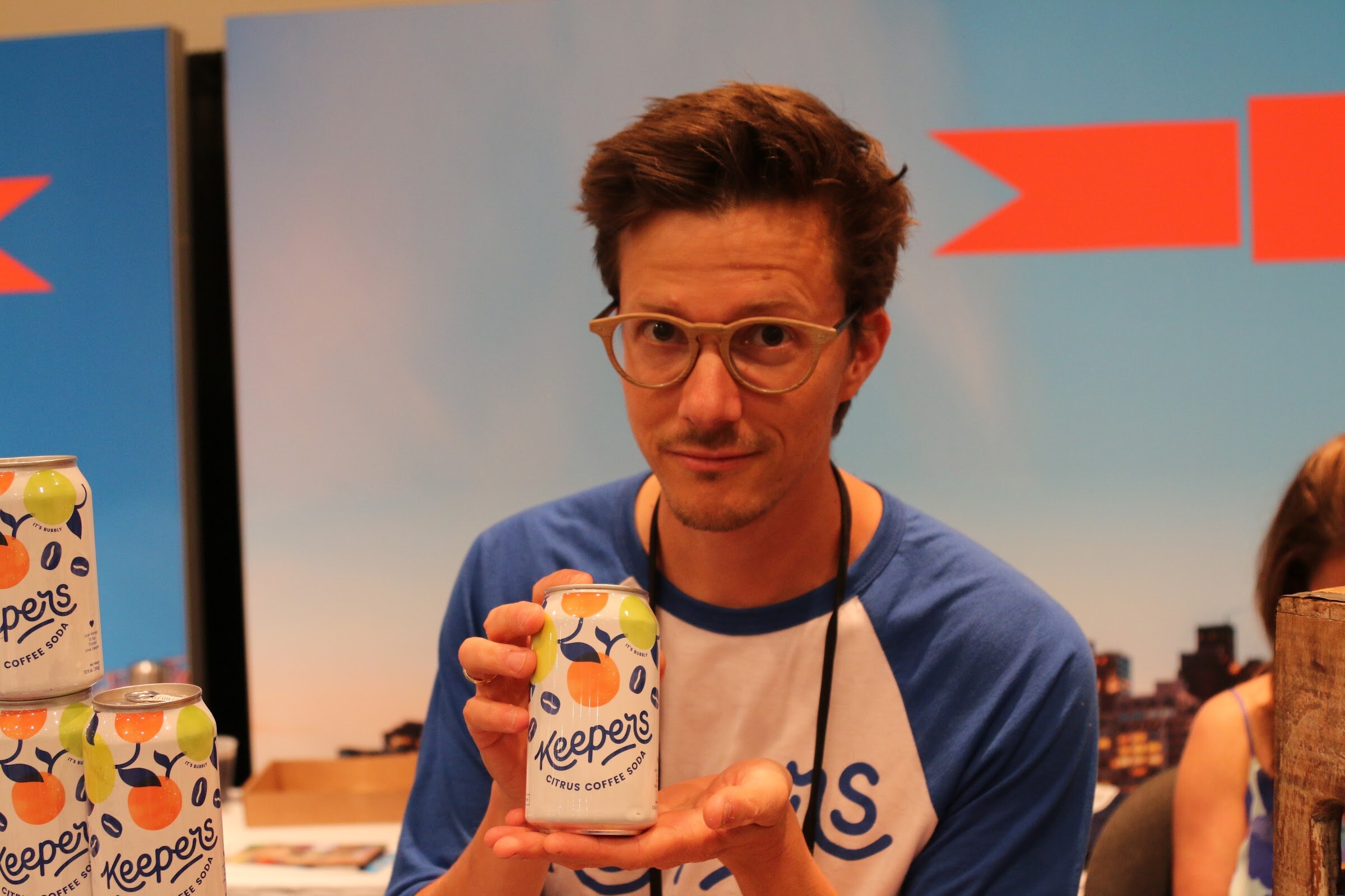 A man with glasses holds a Keepers citrus coffee soda can, with more cans stacked at left and an orange and blue banner behind.