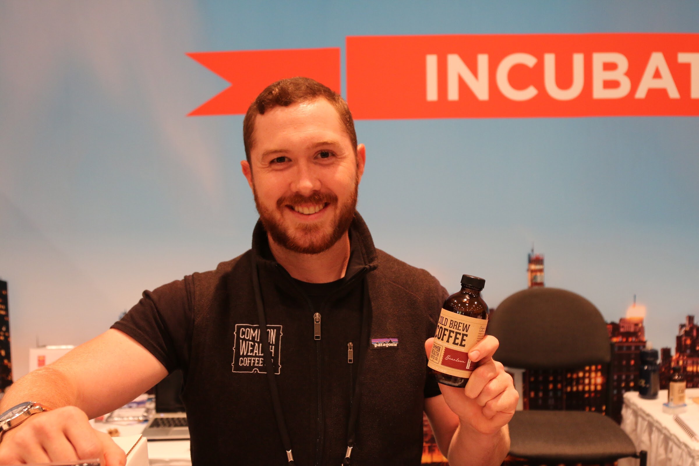 A smiling bearded man in a dark vest holding a small cold brew coffee bottle in front of a blue and orange backdrop.