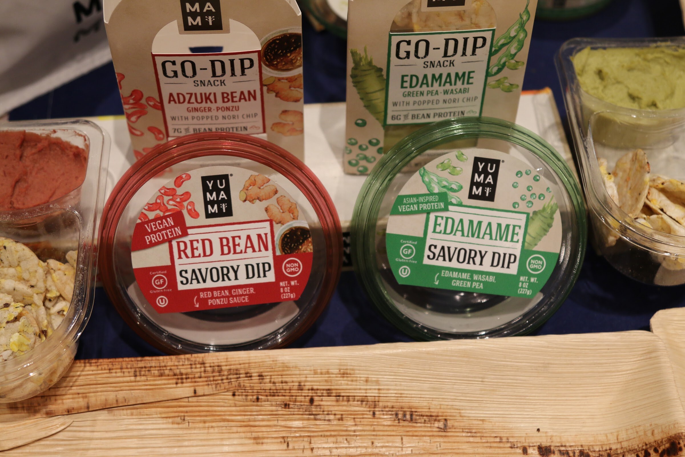 Two tubs of Yumami savory dip, red bean and edamame, in front of Go-Dip snack boxes on a leaf-shaped plate.