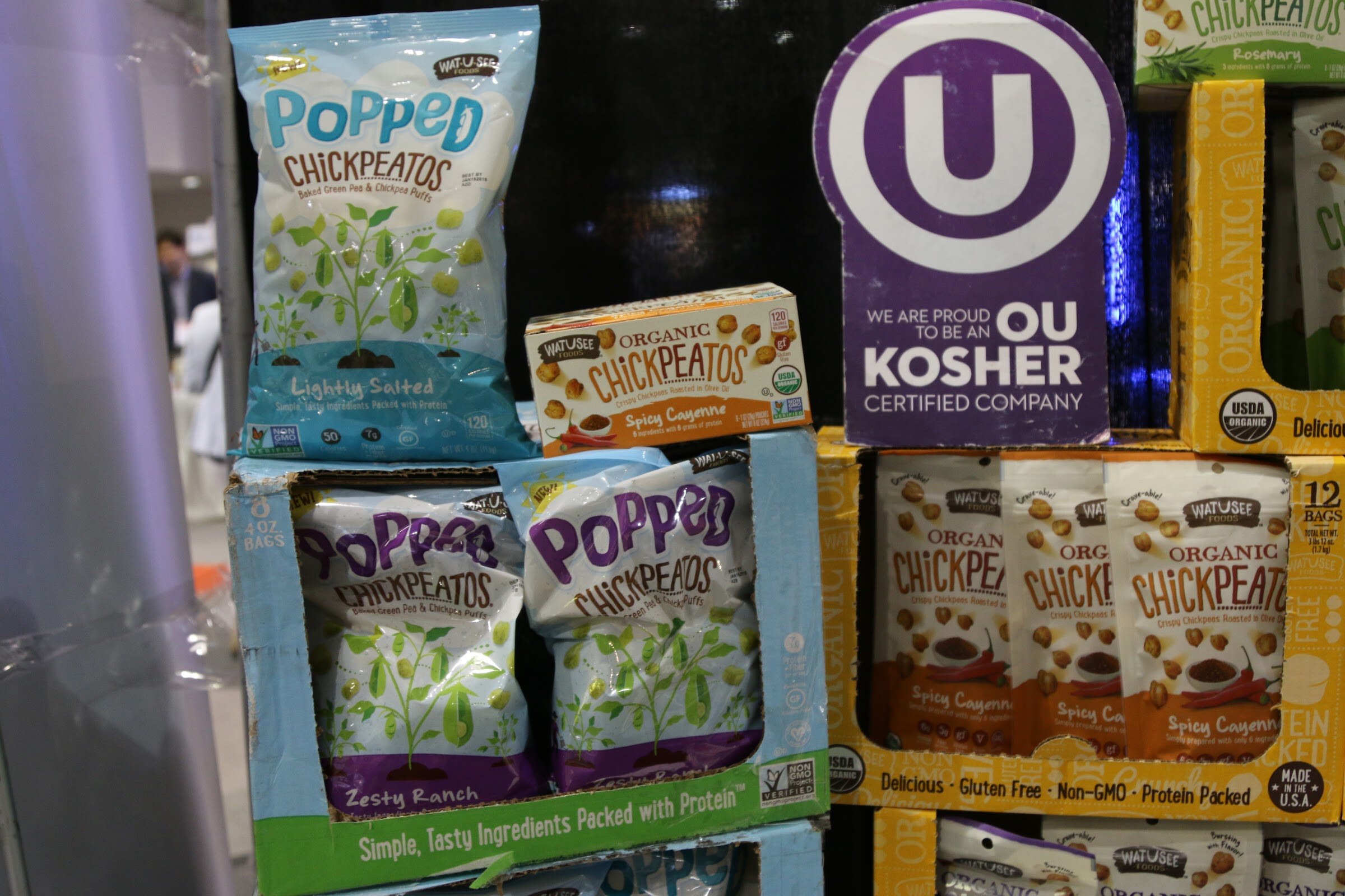 Stacked blue and yellow snack cartons of Popped Chickpeatos with an OU Kosher Certified Company sign.
