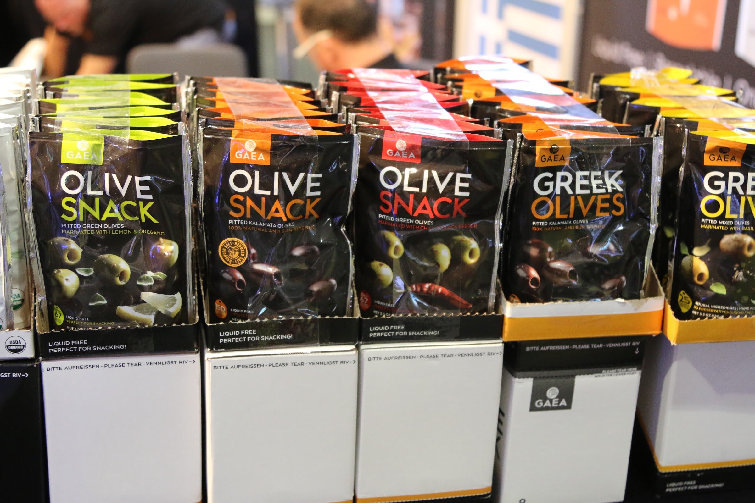 Rows of black Gaea olive snack pouches with green, orange and red labels in white display boxes.