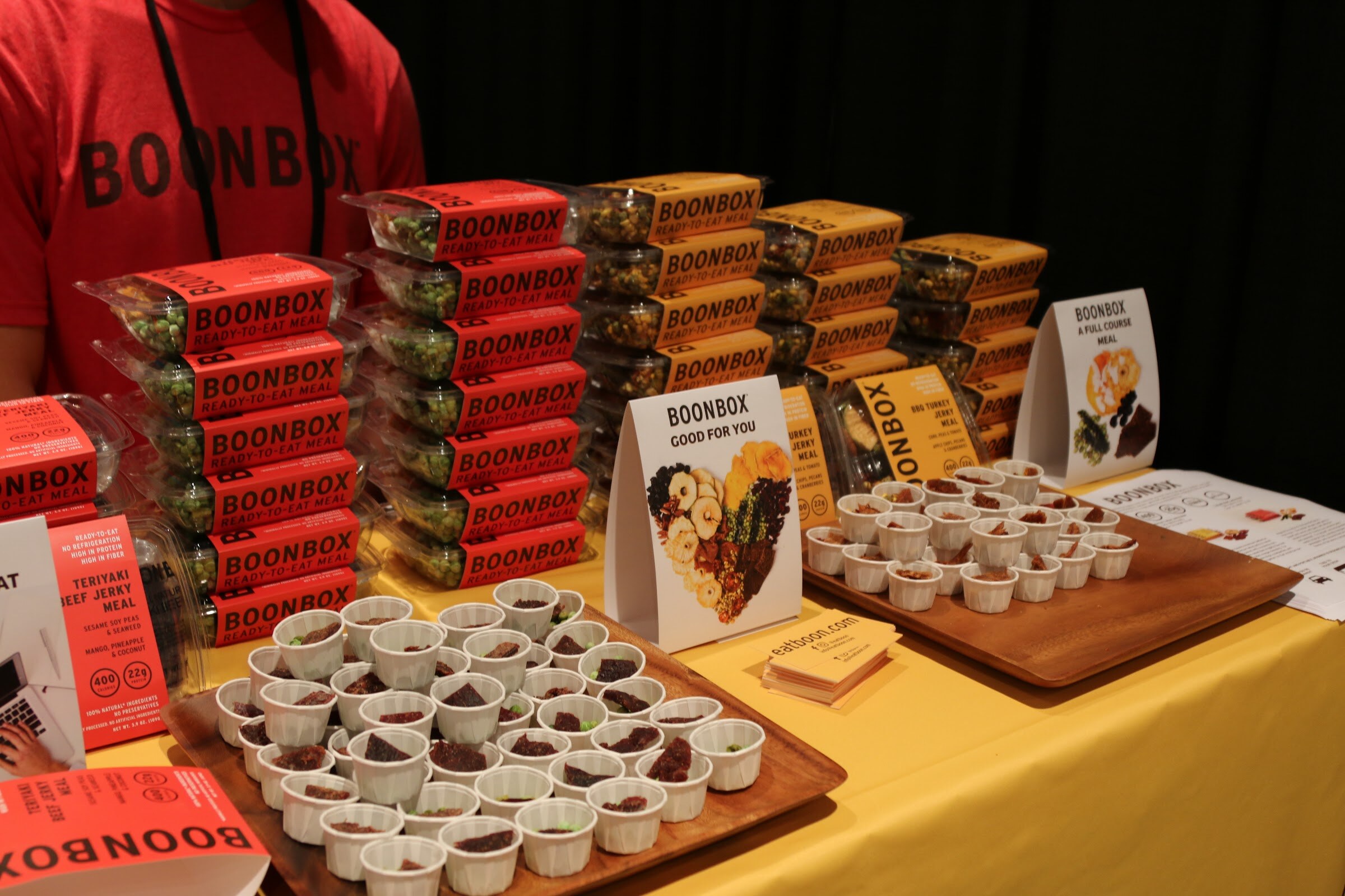 Stacked red and yellow Boonbox ready-to-eat meals behind trays of small paper sample cups on a yellow tablecloth.
