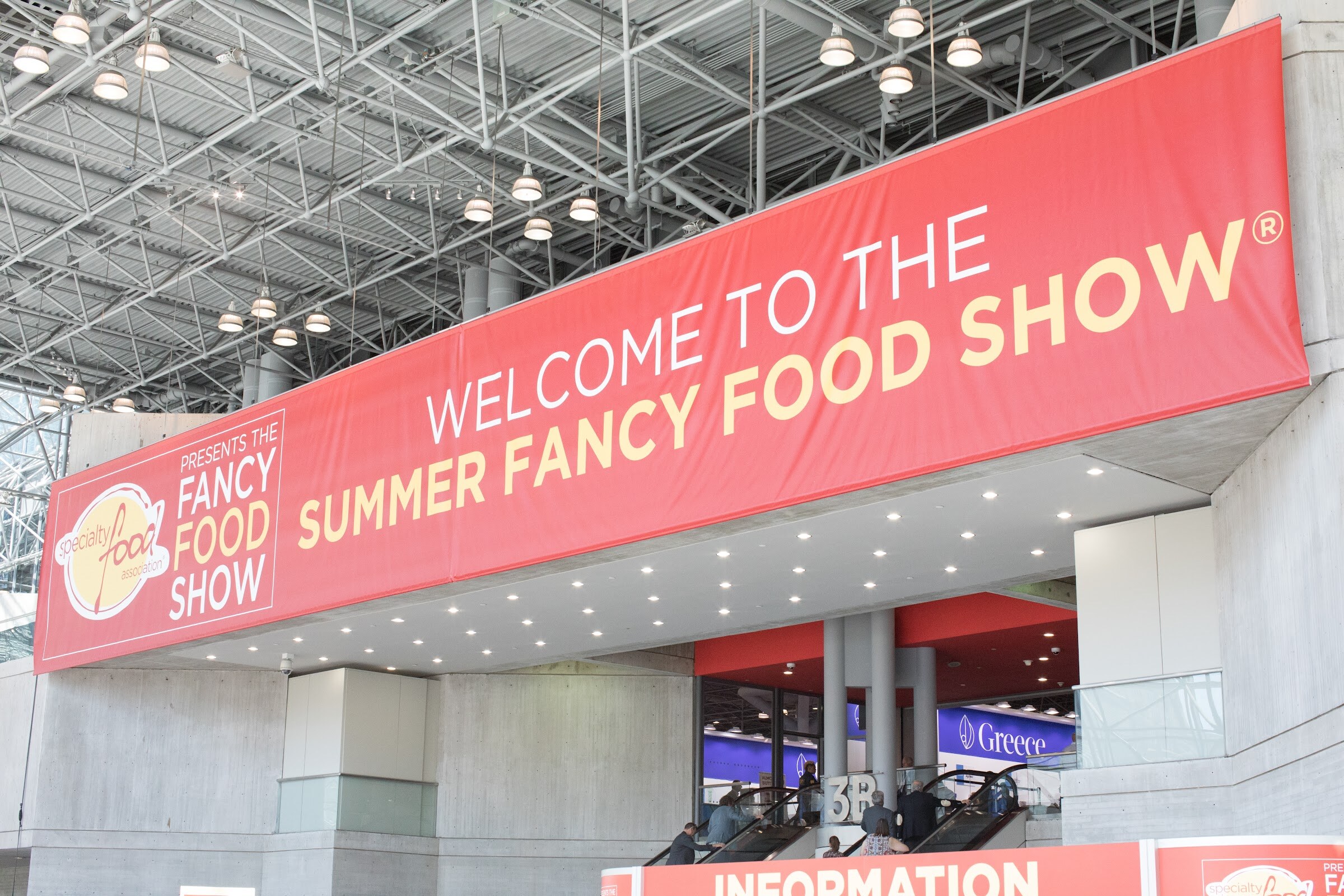 A red banner reading Welcome to the Summer Fancy Food Show with the Specialty Food Association logo above an escalator and information desk.
