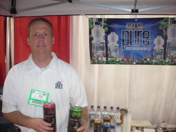 A man in a white Aspen Pure polo with an exhibitor badge holds two canned green tea drinks in front of an Aspen Pure Rocky Mountain Water banner.