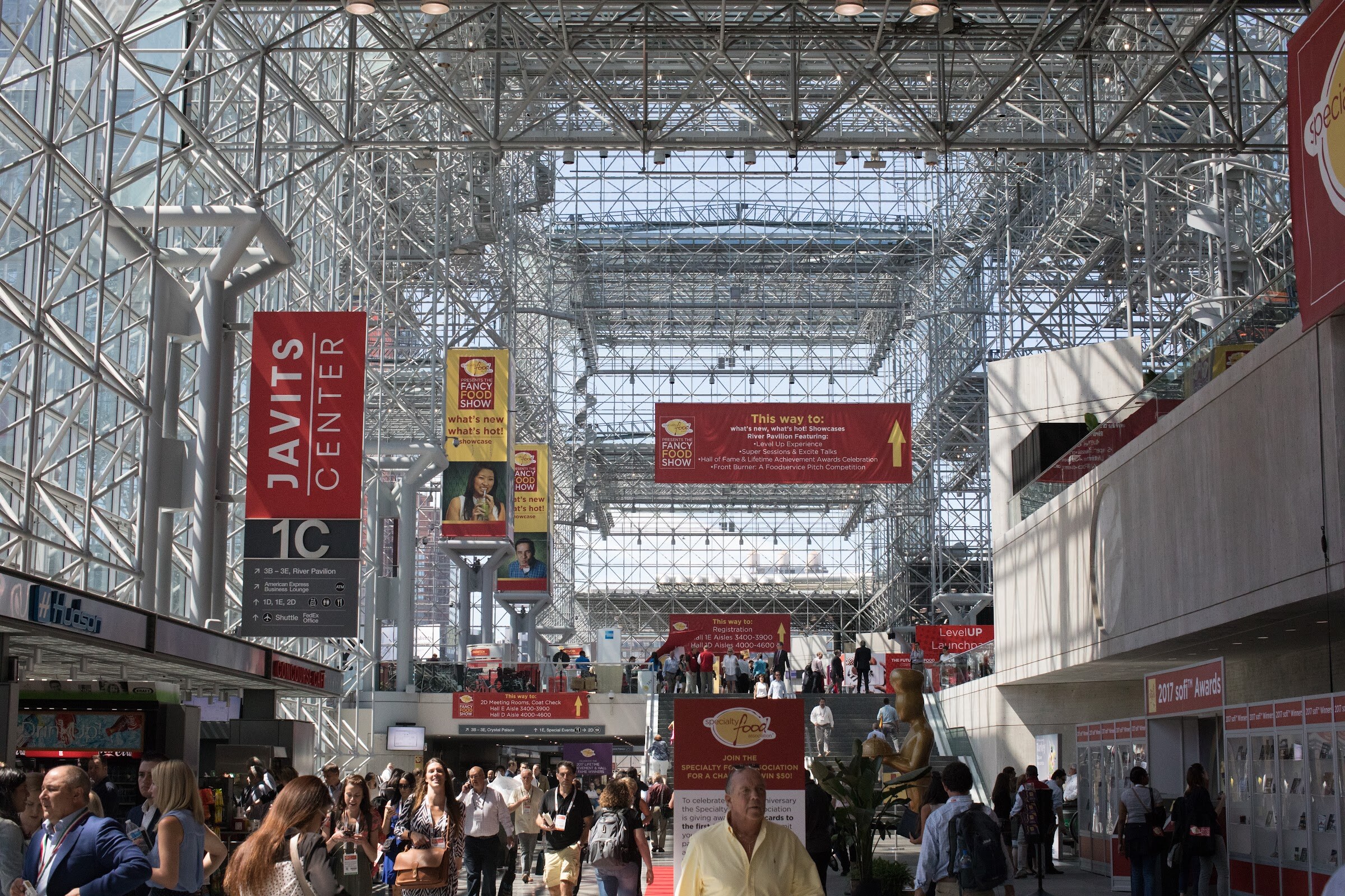 A crowded convention center atrium with Javits Center banners and Fancy Food Show signs under a steel and glass roof.