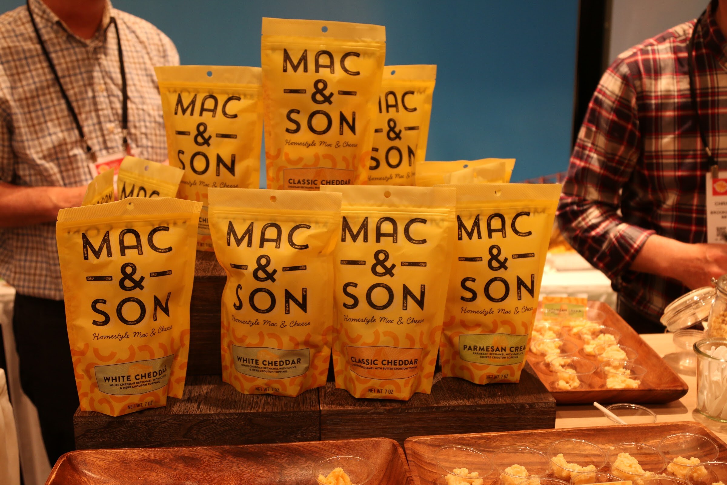 Yellow pouches of Mac & Son mac and cheese stacked on wooden risers with sample trays below and two men in plaid shirts behind.
