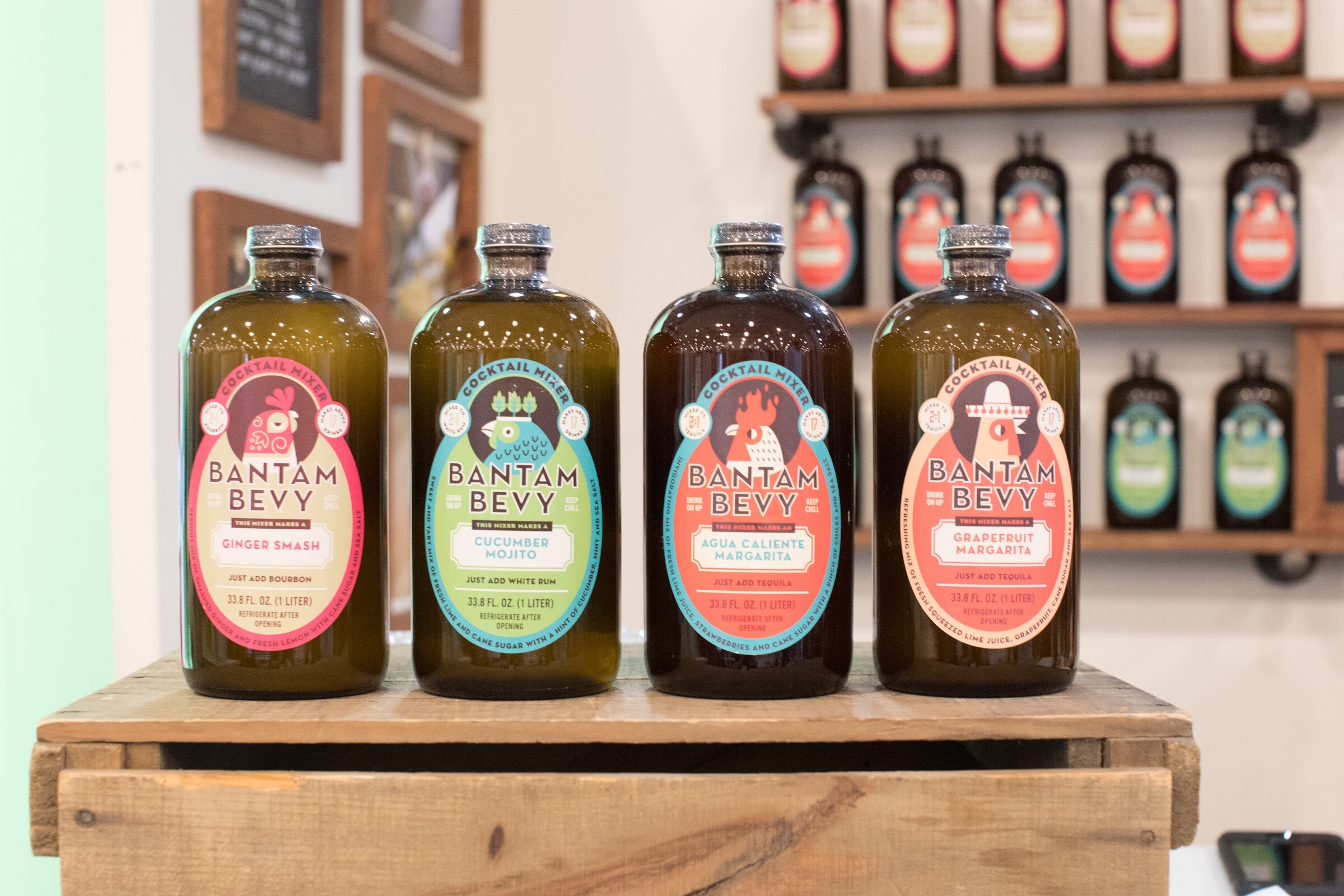 Four dark glass Bantam Bevy bottles with colorful rooster labels on a wood crate, with more bottles on a shelf behind.