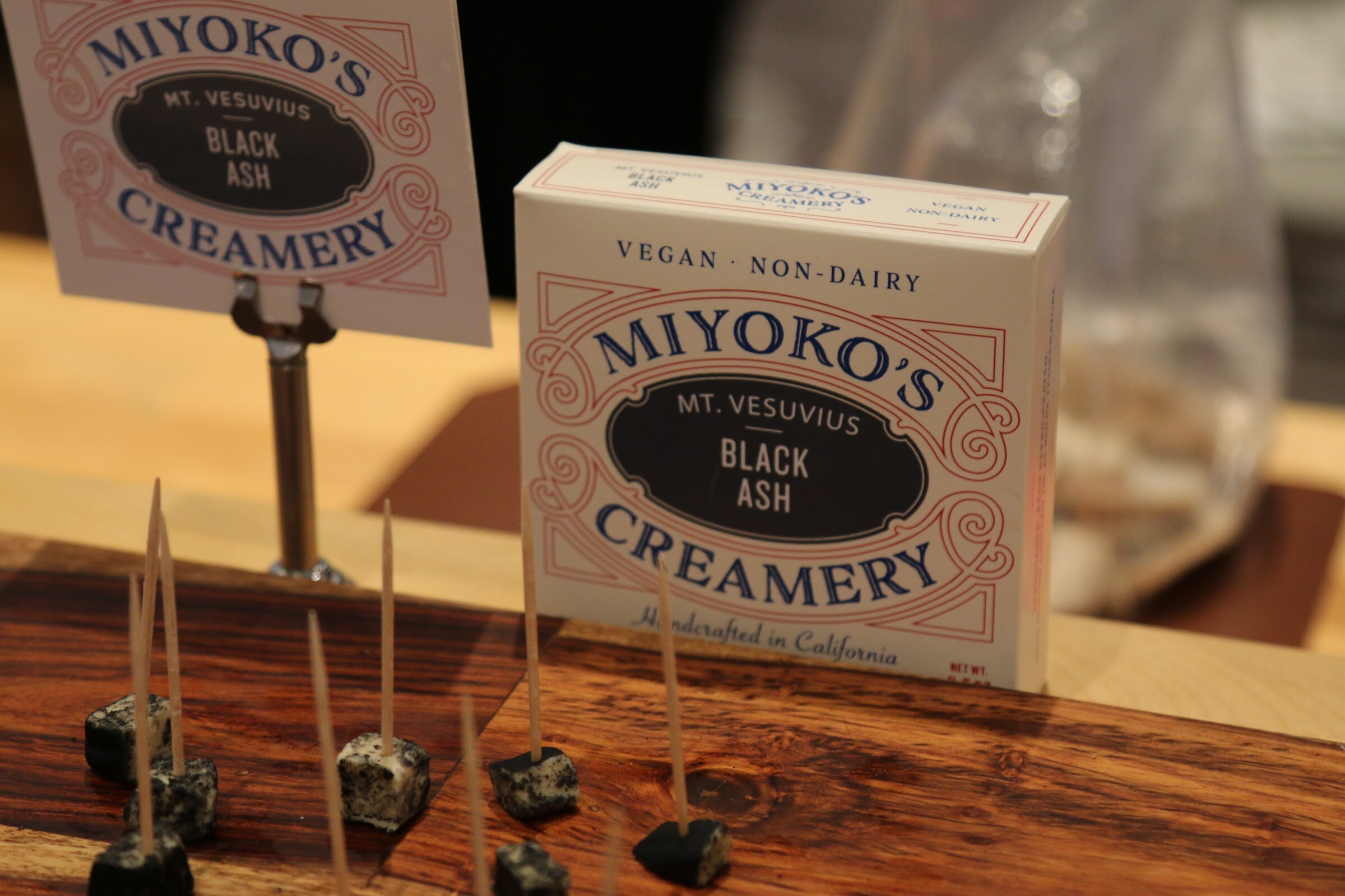 A Miyoko's Creamery Mt. Vesuvius Black Ash box and sign behind small cubes of cheese on toothpicks.