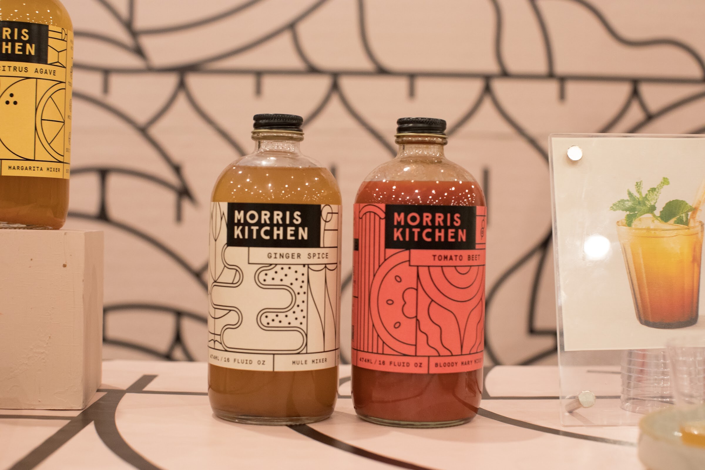 Two bottles of Morris Kitchen mixer with black labels in front of a pink wall with line patterns.