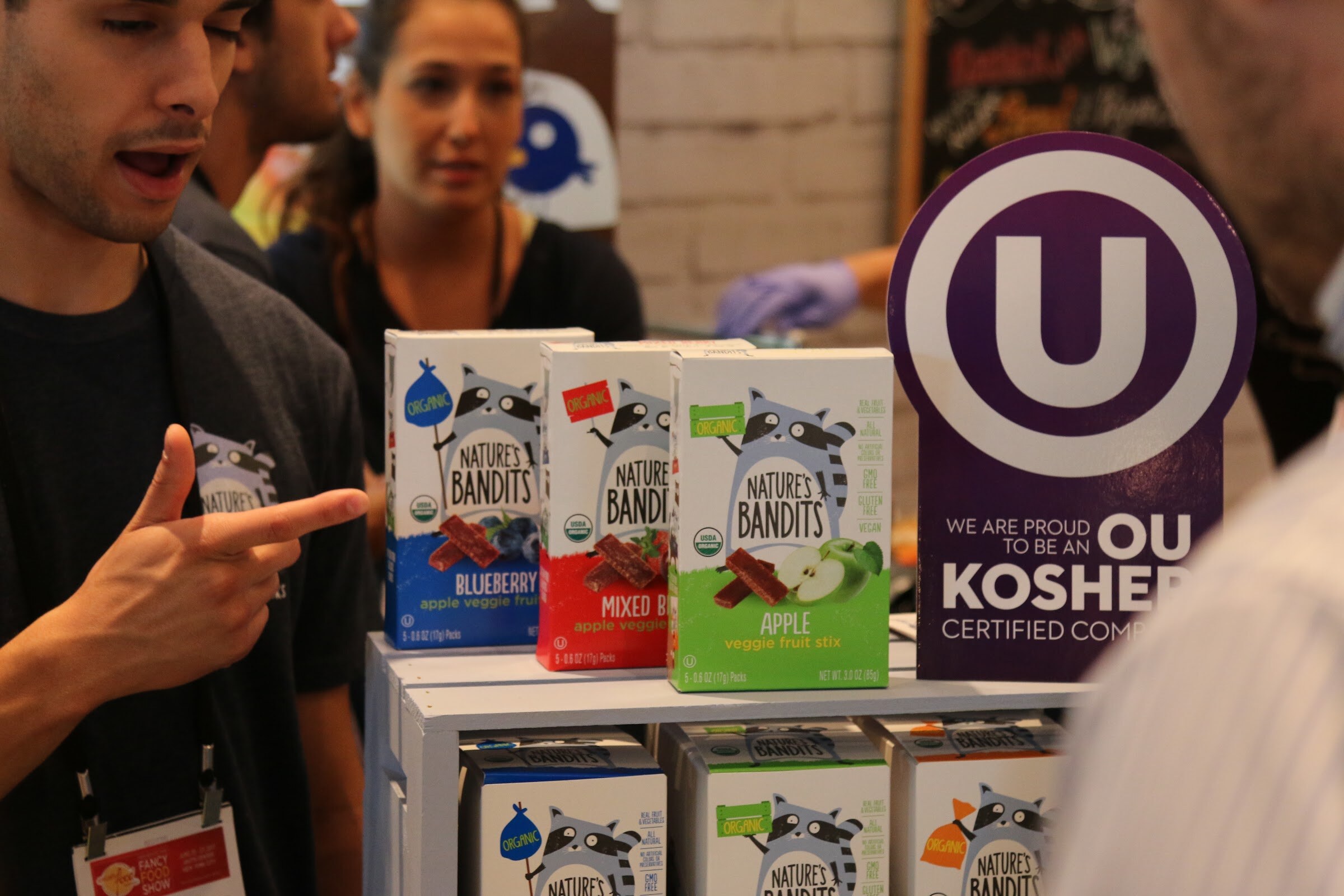A man in a dark shirt points while talking beside boxes of Nature's Bandits fruit sticks and a purple OU Kosher sign.