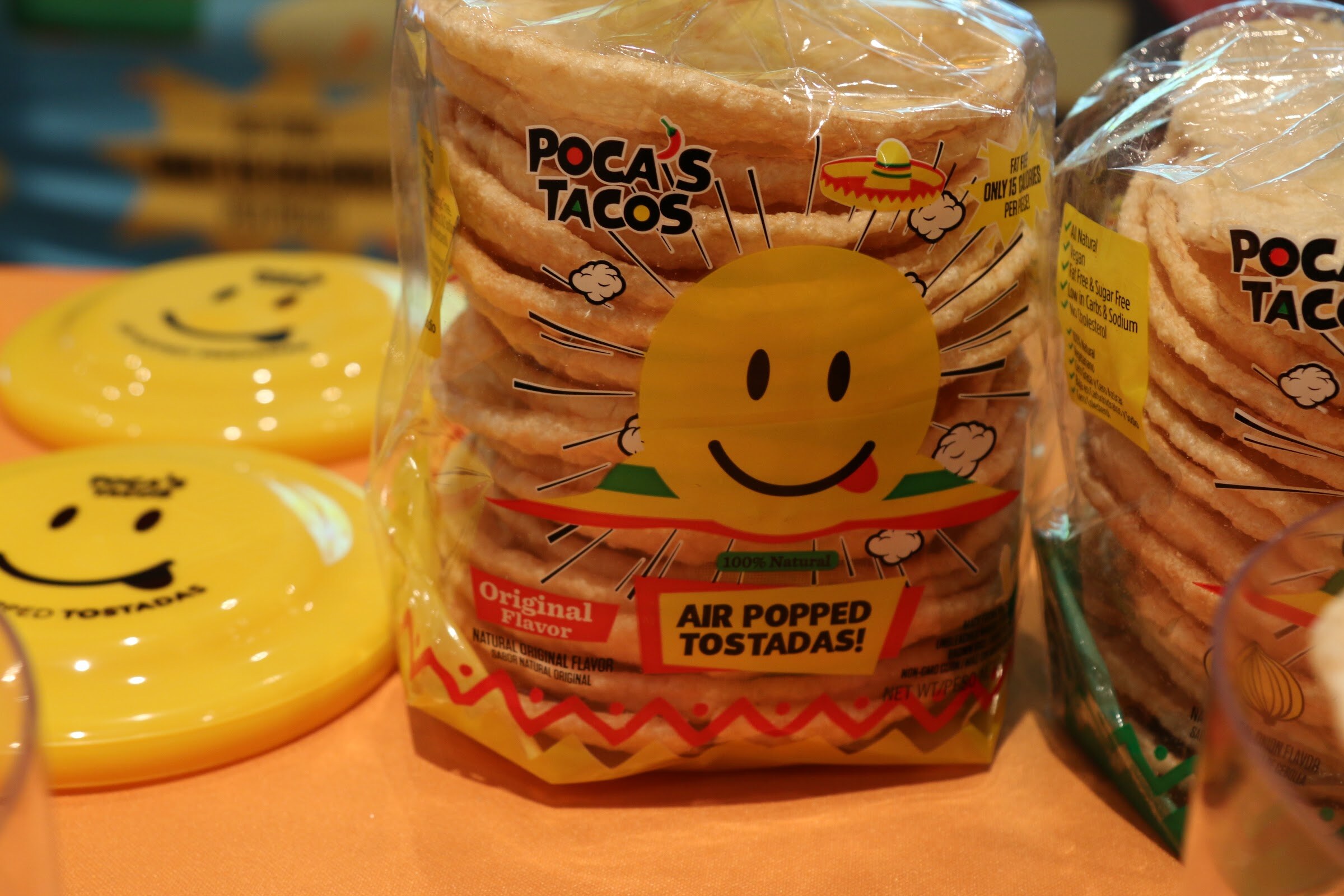 A clear bag of Poca's Tacos tostadas with a smiling sun logo, with yellow smiley-face discs on an orange tablecloth.