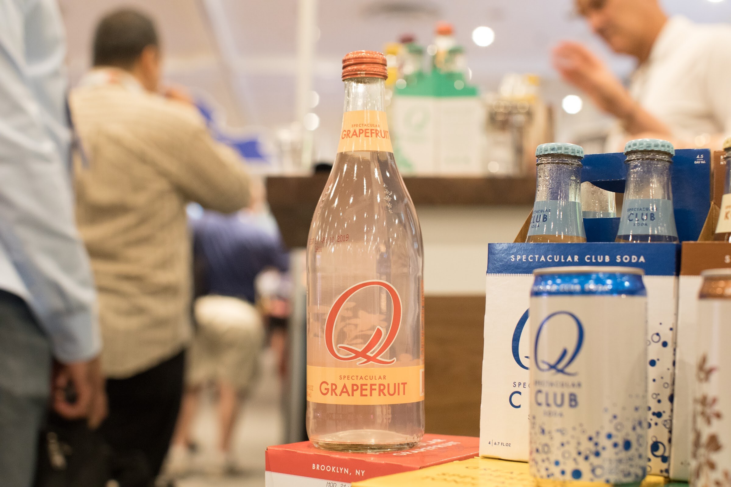 An orange-capped Q Grapefruit soda bottle beside blue Spectacular Club soda cans, with blurred people in the background.