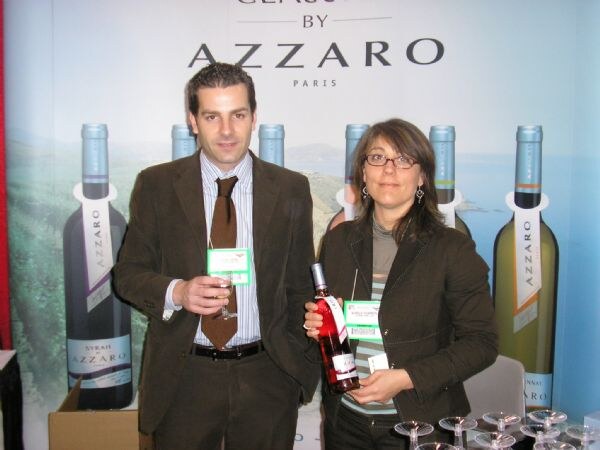 A man in a brown suit holding a glass and a woman with glasses holding a bottle of rosé stand before a backdrop of Azzaro Paris wine bottles.