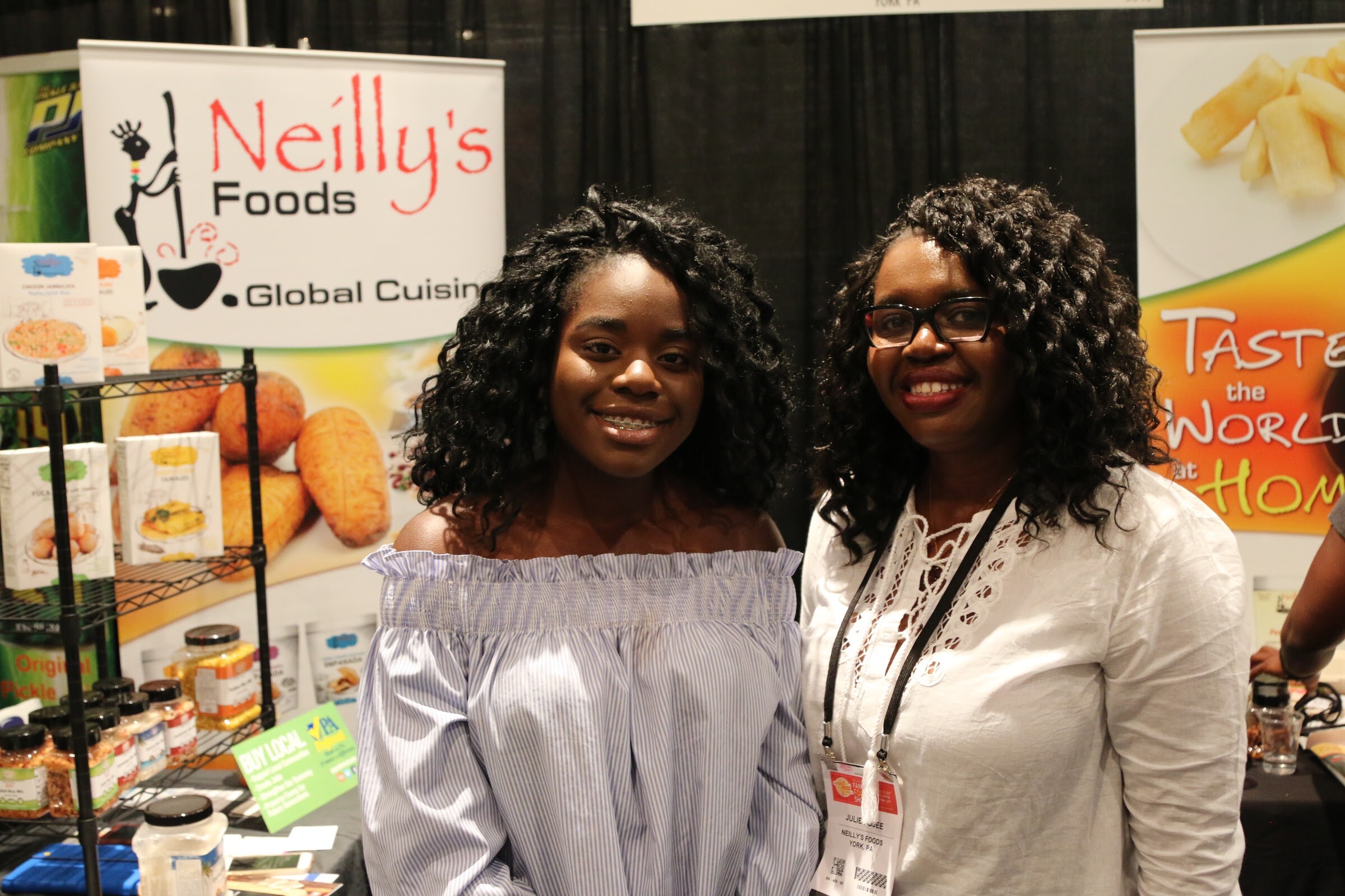 Two smiling women in front of a banner reading Neilly's Foods Global Cuisine, with product boxes on shelves.