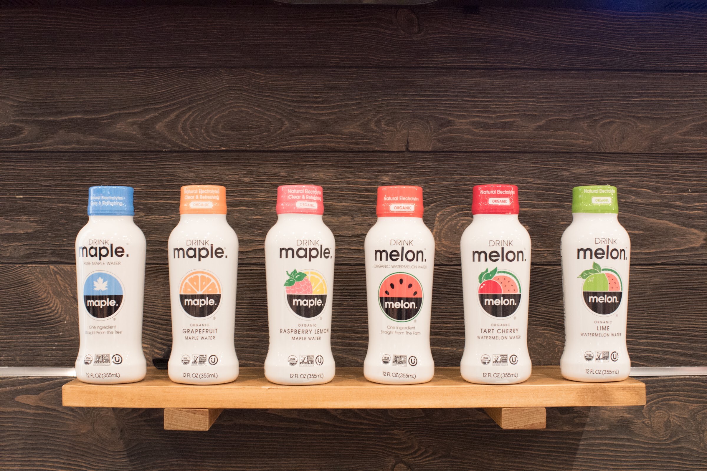 Six white bottles of maple water and watermelon water on a wooden shelf in front of dark wood planks.