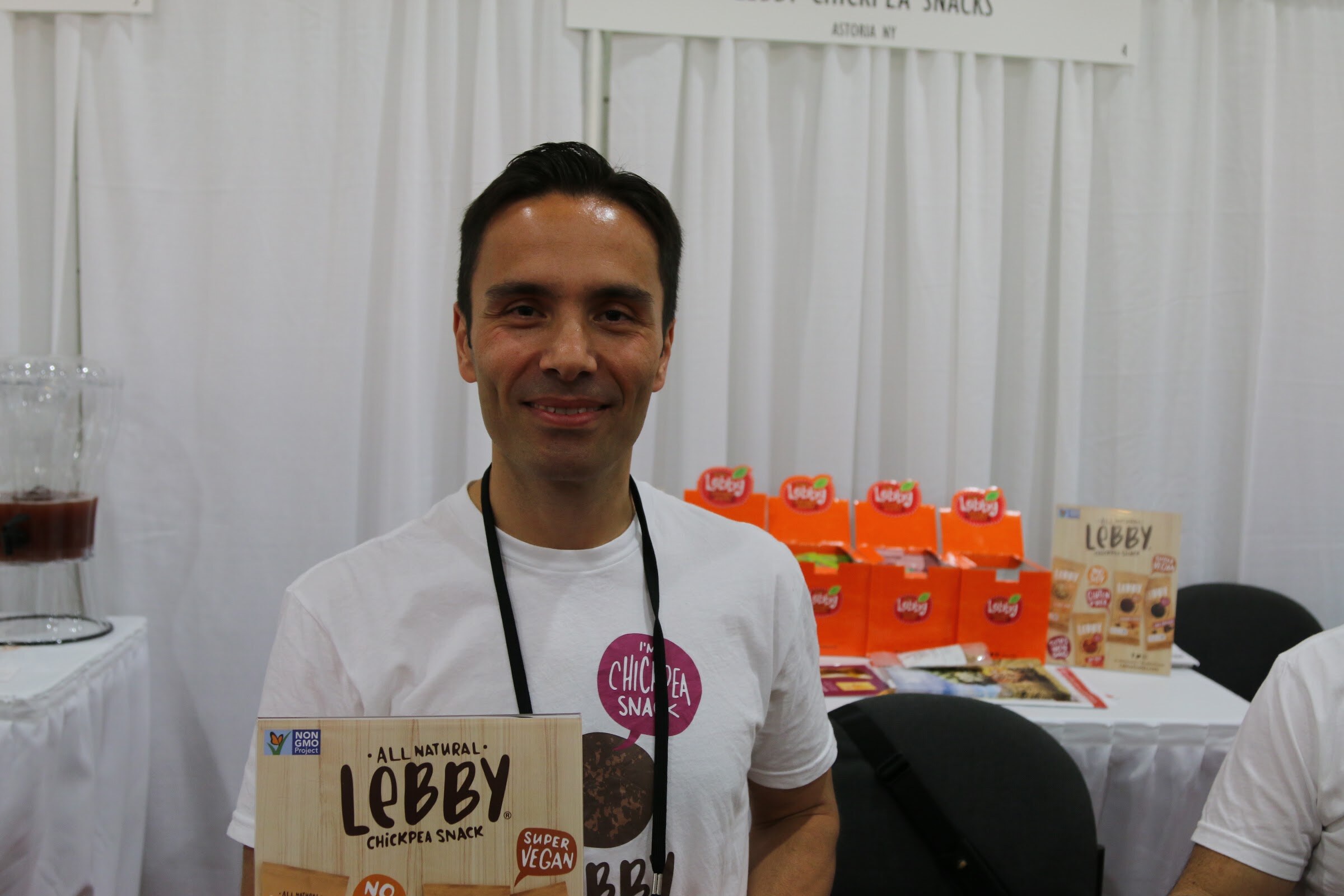 A man in a white t-shirt holding a box of Lebby chickpea snacks, with orange Lebby boxes on the table behind him.