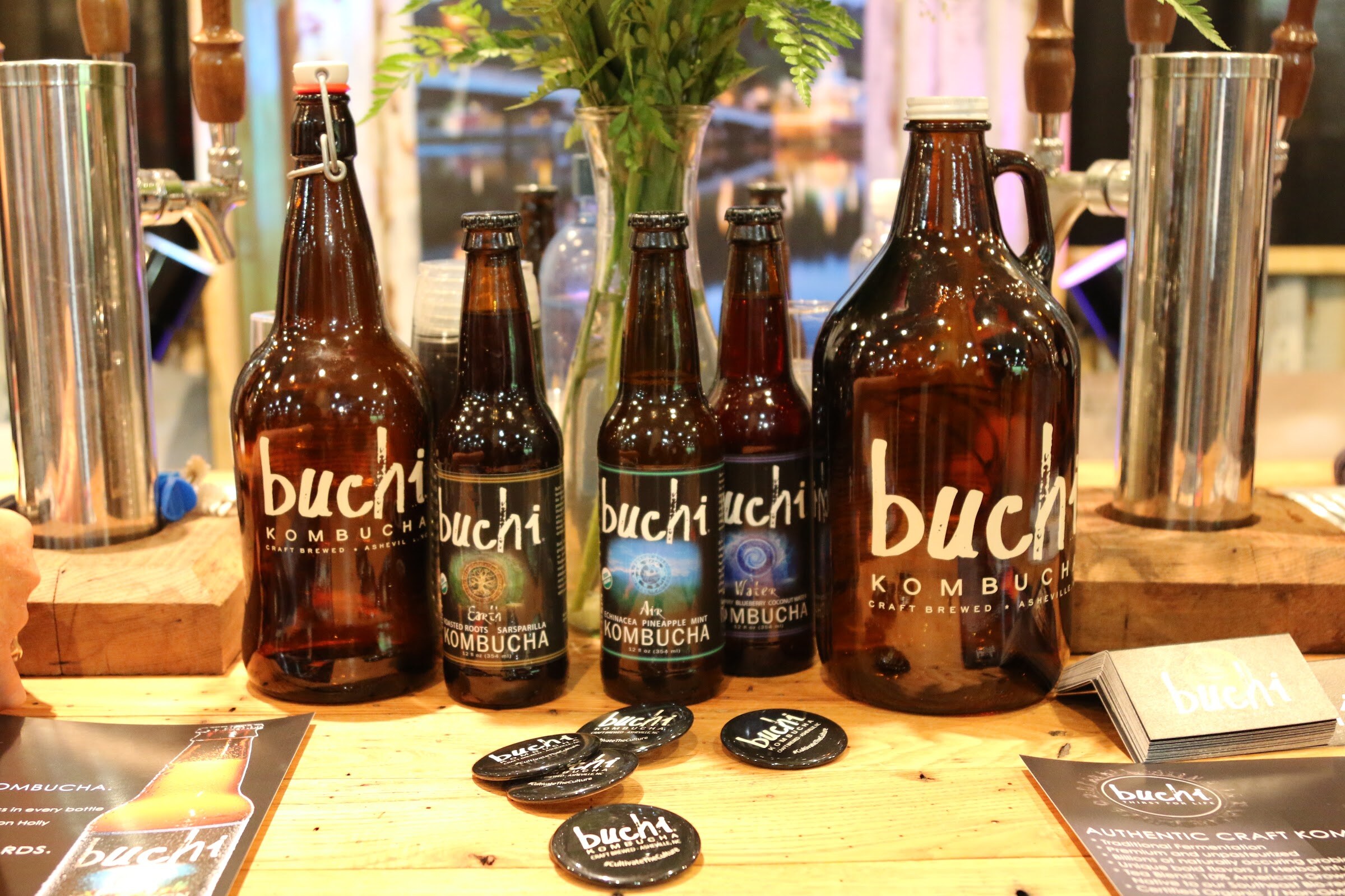 Brown Buchi kombucha bottles and two growlers on a wooden counter with tap handles and buttons.