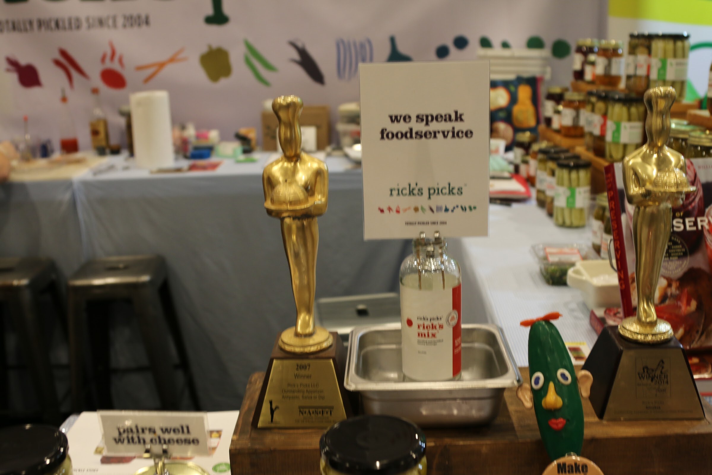 Two gold chef trophies beside a Rick's Picks bottle and a sign saying we speak foodservice.
