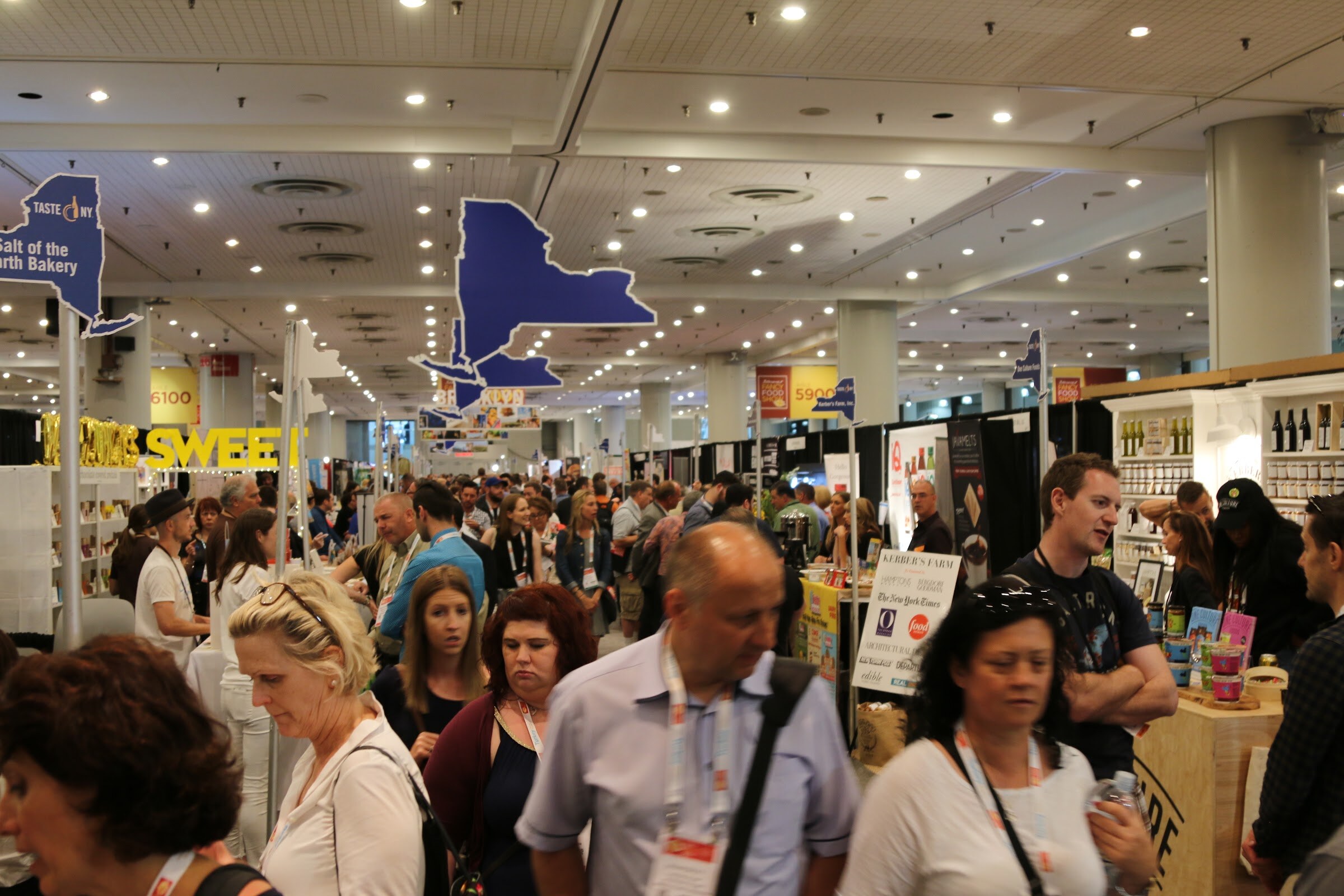 A dense crowd of badge-wearing attendees on a convention floor beneath ceiling lights and blue New York state shapes.