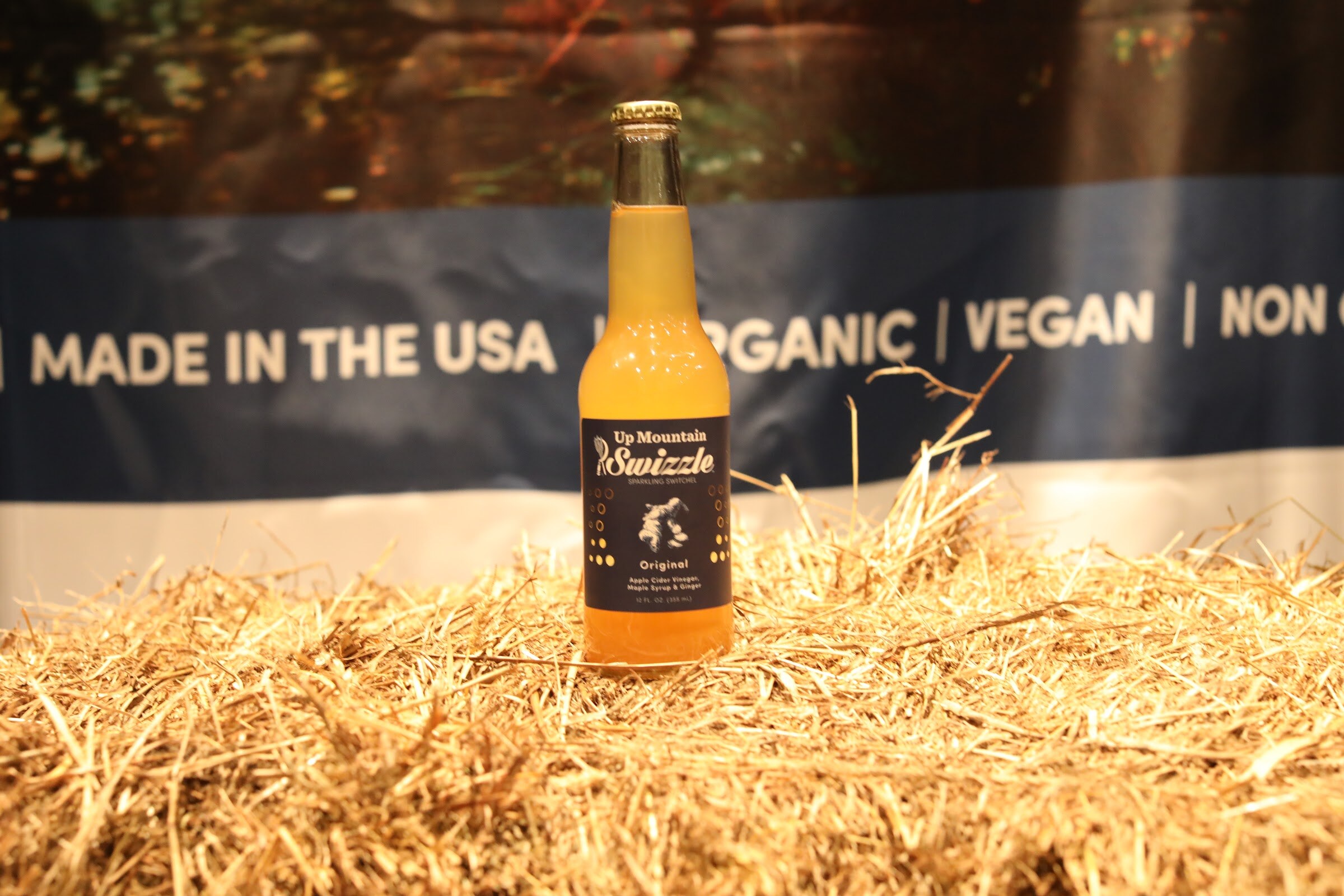 A bottle of Up Mountain Swizzle on a bed of straw in front of a blue banner.