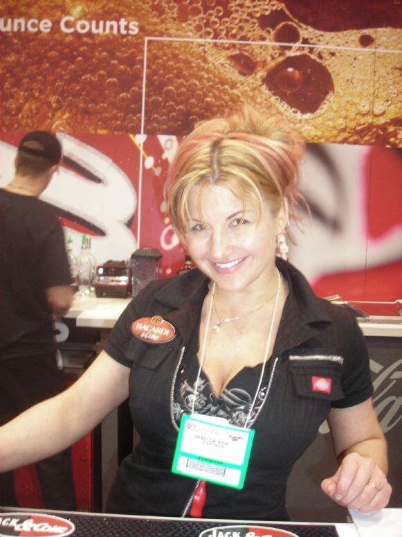 A smiling woman in a black Bacardi & Coke shirt with an exhibitor badge stands at a bar counter; a person in a cap stands behind her near Coca-Cola branding.