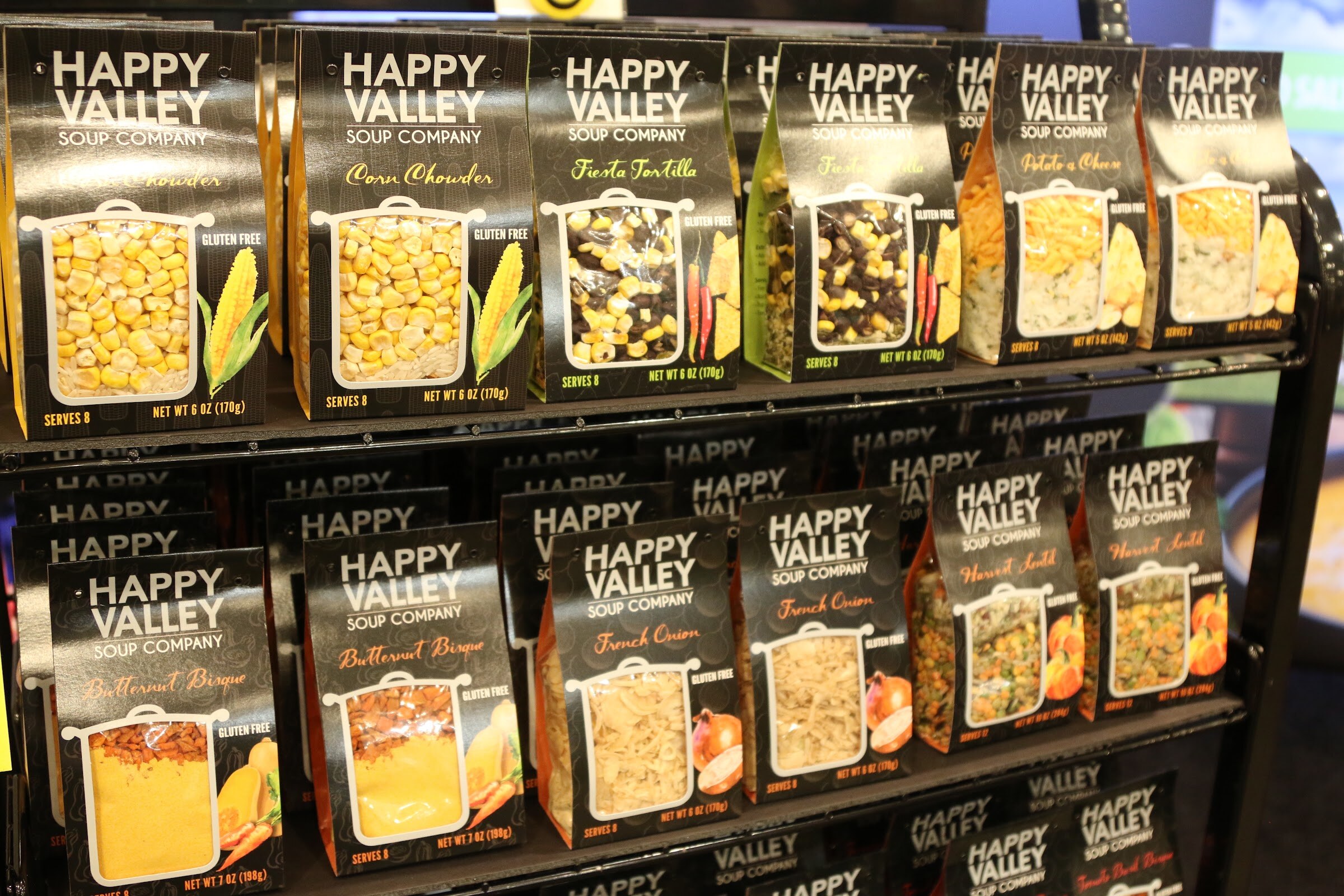 Rows of Happy Valley Soup Company packages with clear windows showing dried soup ingredients on a metal rack.