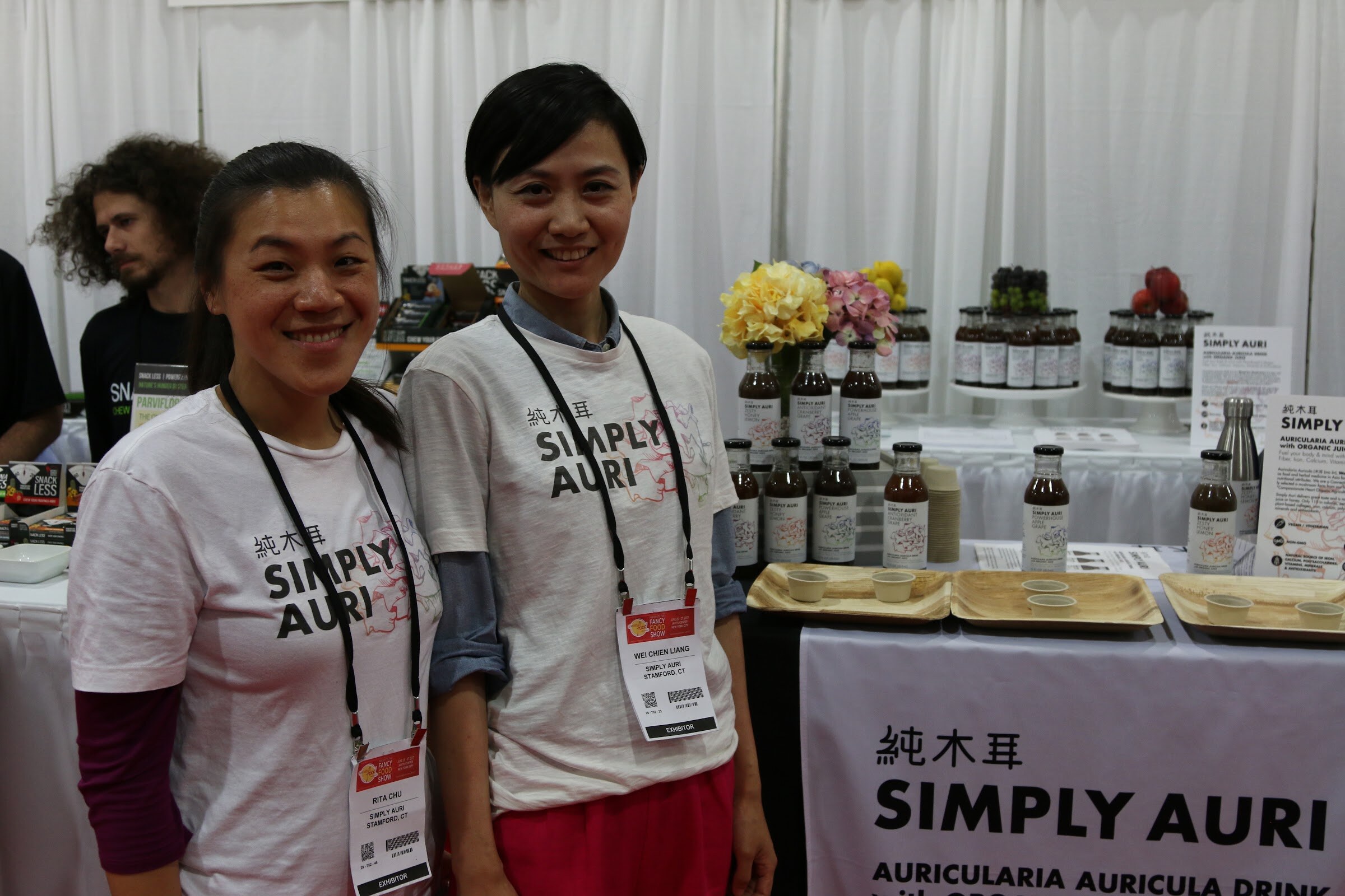 Two smiling women with badges in Simply Auri t-shirts in front of a table of bottles and sample trays.