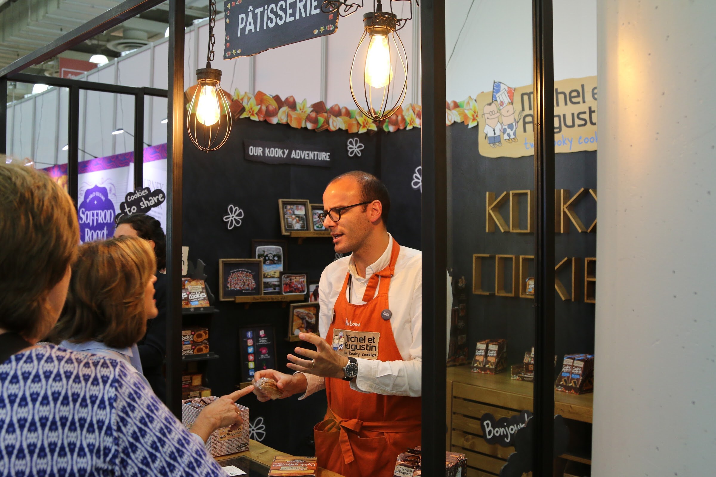 A man in an orange apron gesturing to visitors in front of a dark booth with hanging lights and a Michel et Augustin sign.