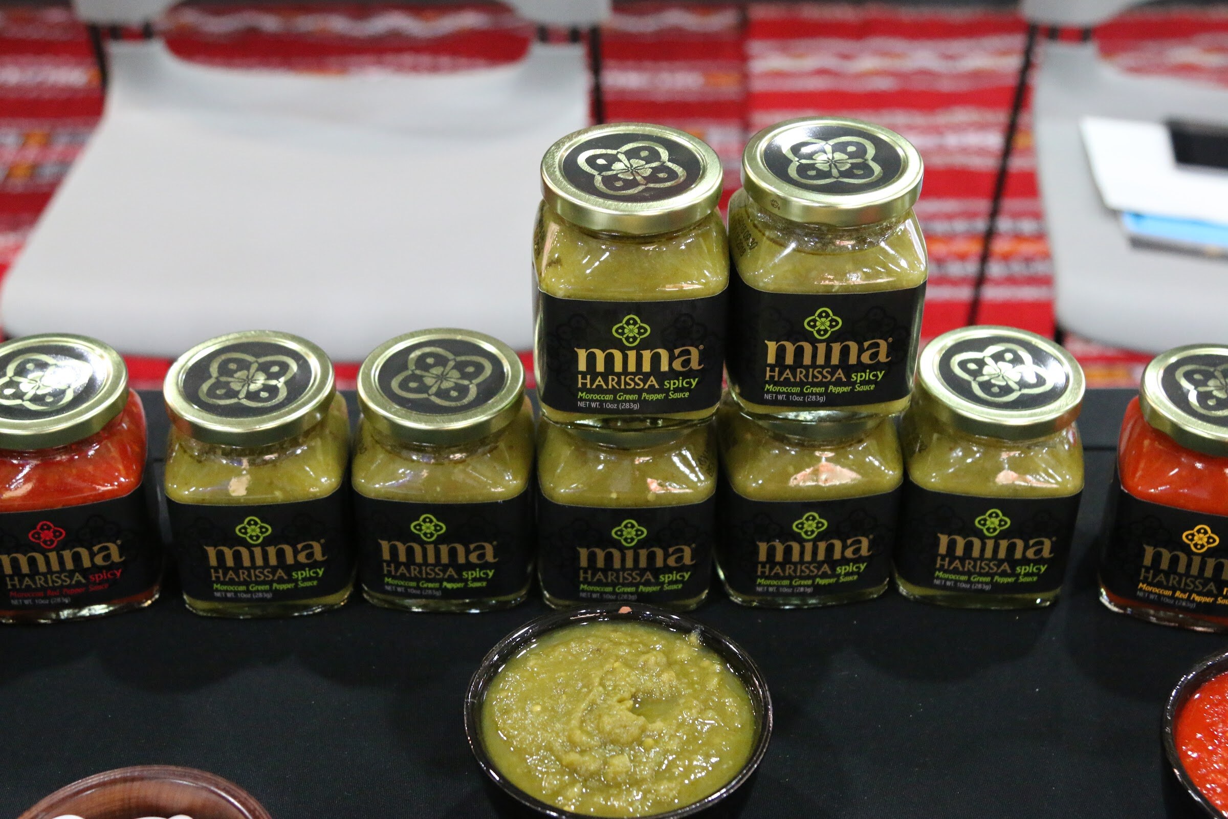 Rows of Mina Harissa sauce jars with black labels on a black table and a bowl of green sauce.