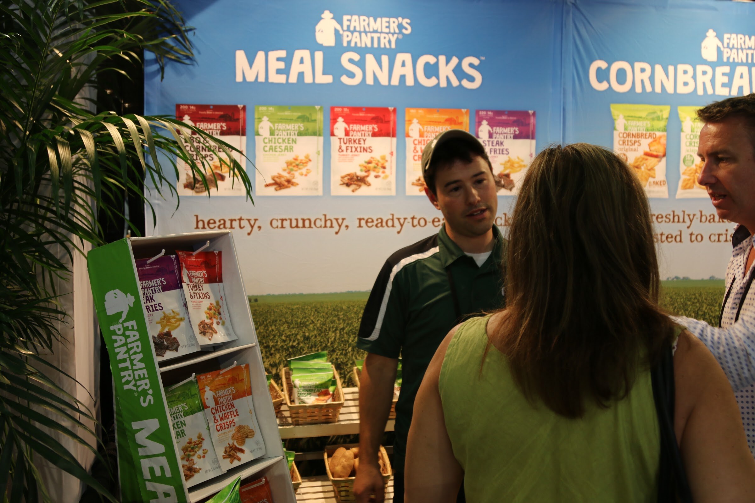 A man in a green polo talks with a woman seen from behind in front of a Farmer's Pantry Meal Snacks banner and display rack.