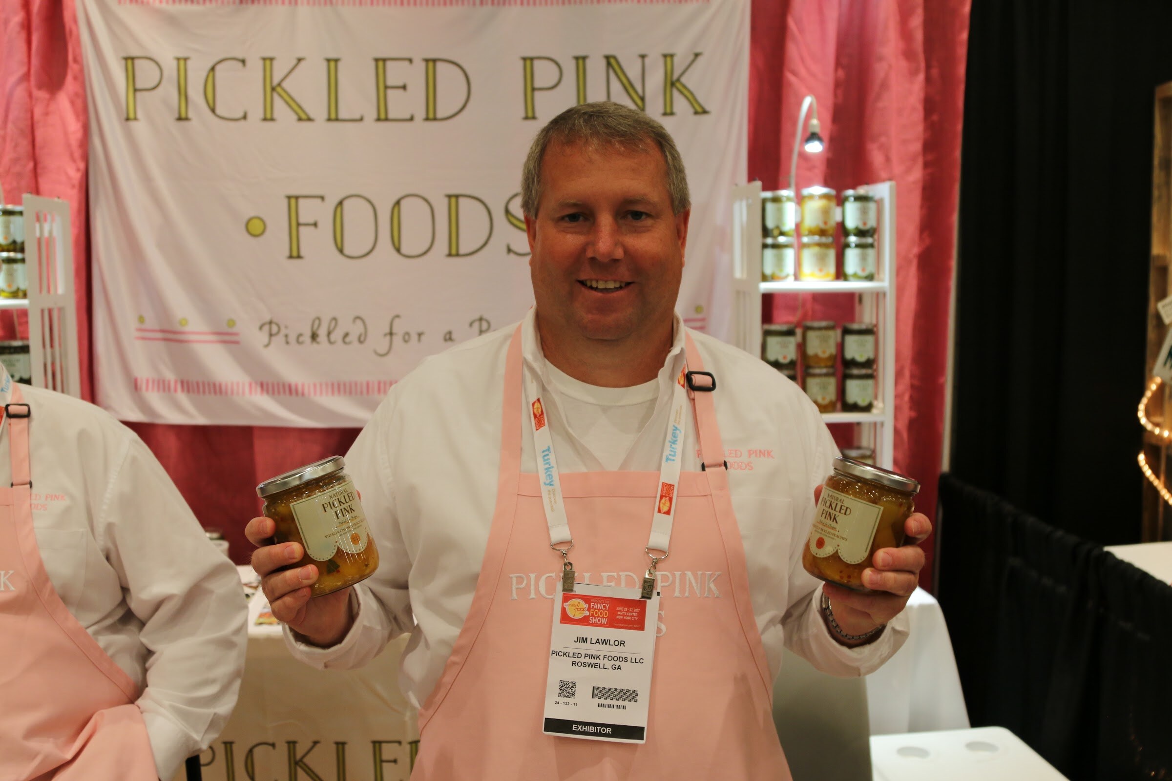 A man in a pink apron with an exhibitor badge holding two jars of pickles under a Pickled Pink Foods banner.